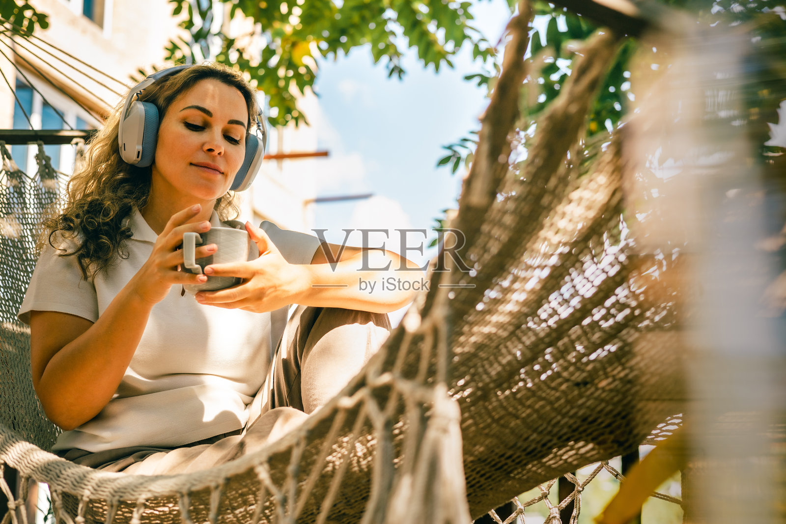戴着耳机、躺在吊床上、在阳光明媚的户外露台上享用热饮的女性照片摄影图片