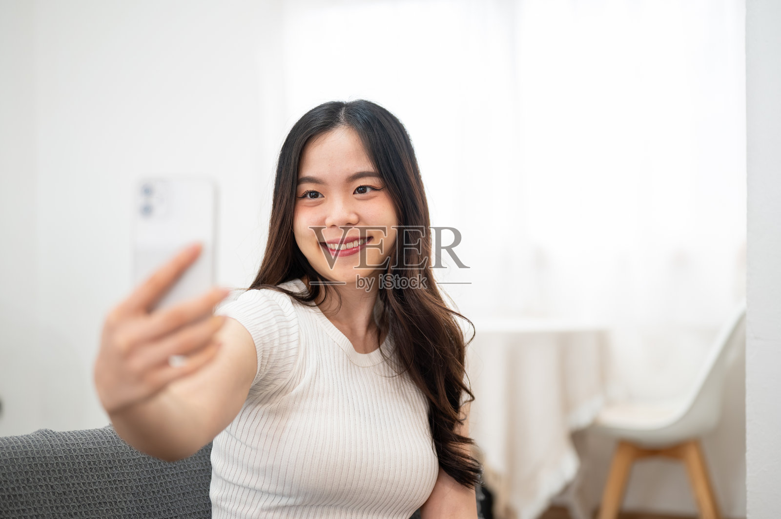 一个快乐的亚洲女子坐在白色客厅的沙发上，手持智能手机自拍照片摄影图片