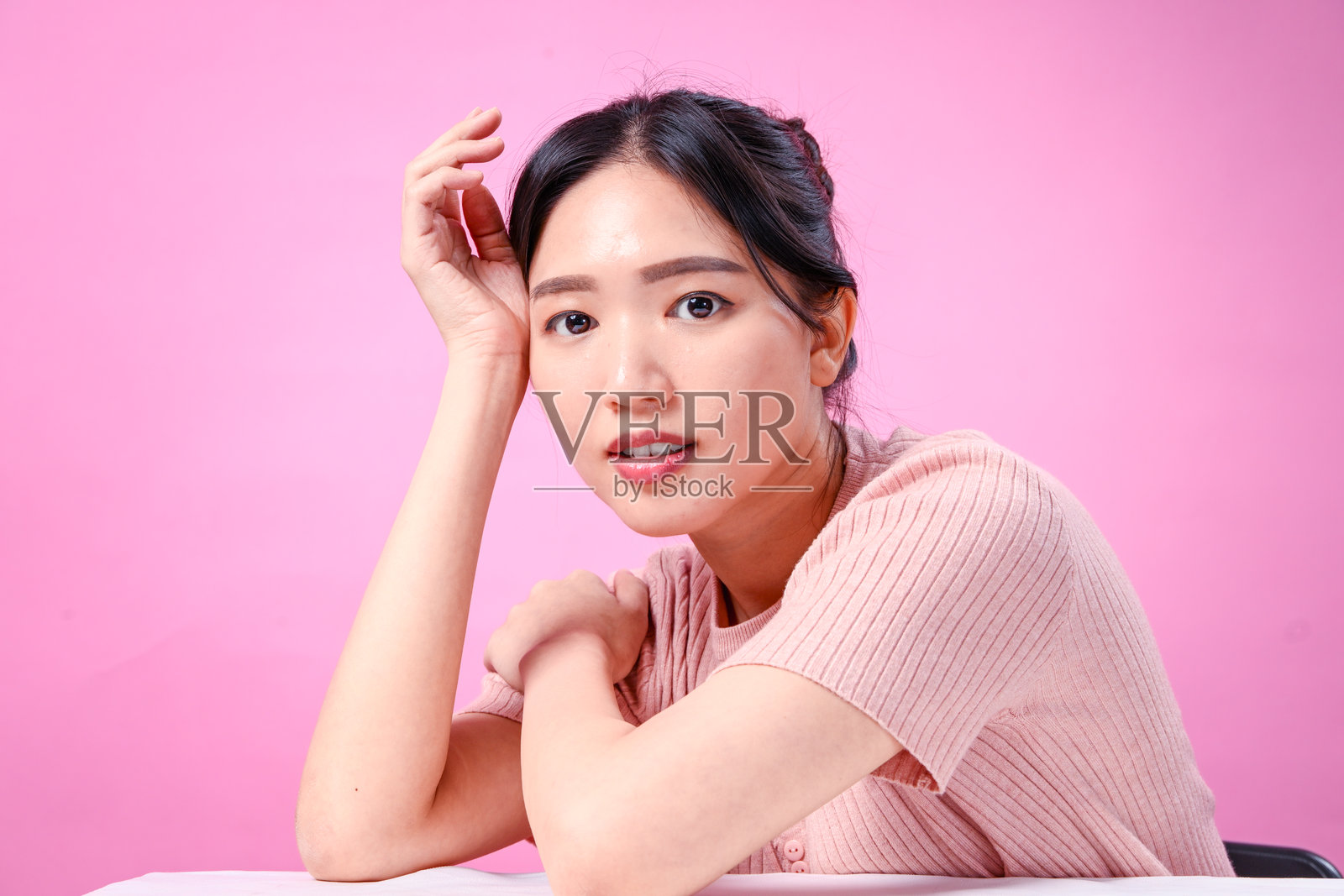 一位留着发髻黑发的年轻亚洲女性，身穿罗纹粉色短袖上衣，单手扶头优雅地摆姿势，在柔和的粉色背景前展现美丽照片摄影图片