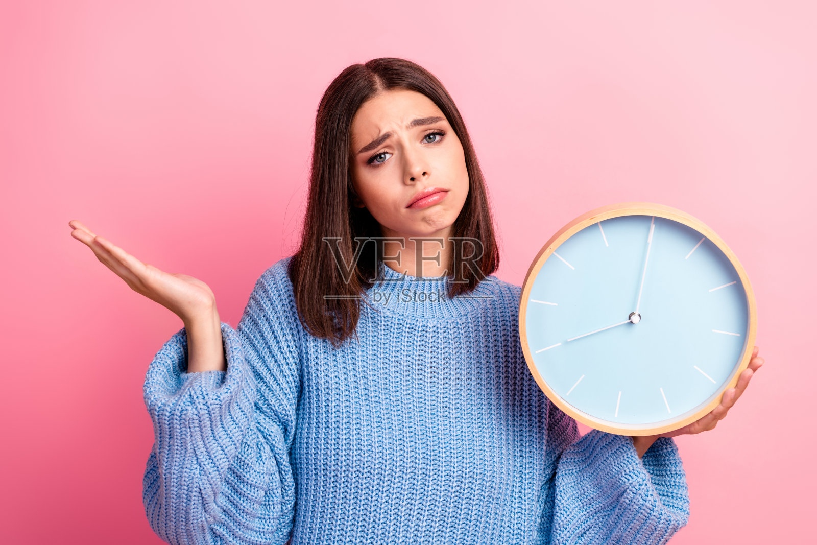 一位穿着蓝色毛衣的年轻女子，手持时钟，背景为粉色，她脸上带着困惑的表情，展现了休闲的生活方式照片摄影图片