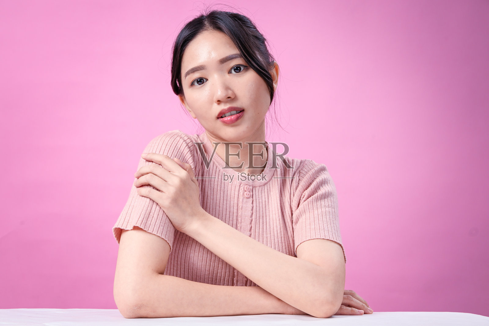 一位留着盘发的年轻亚洲女性，身穿罗纹粉色短袖上衣，优雅地将手搭在肩上，在柔和的粉色背景下展现美丽。照片摄影图片