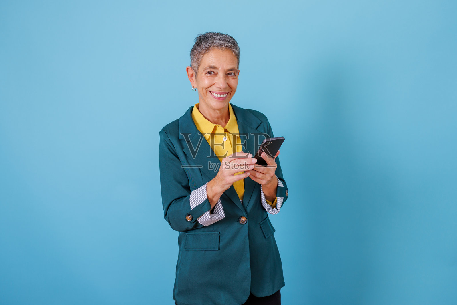 一位年长的女商人正在使用智能手机，她在蓝色的背景下微笑着发短信，享受着现代科技和通讯的便利。照片摄影图片