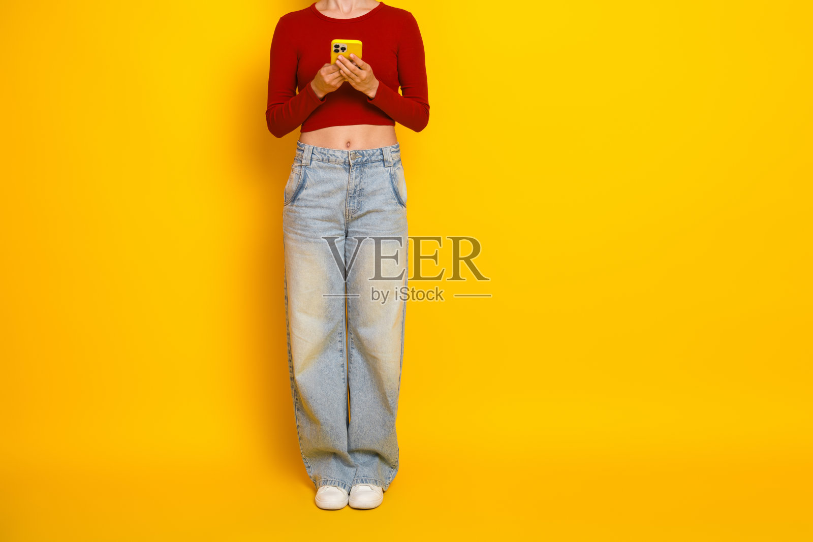身穿红色露脐上衣和宽松牛仔裤的年轻女子，手持黄色智能手机，背景为亮黄色，适用于休闲时尚和广告宣传。照片摄影图片