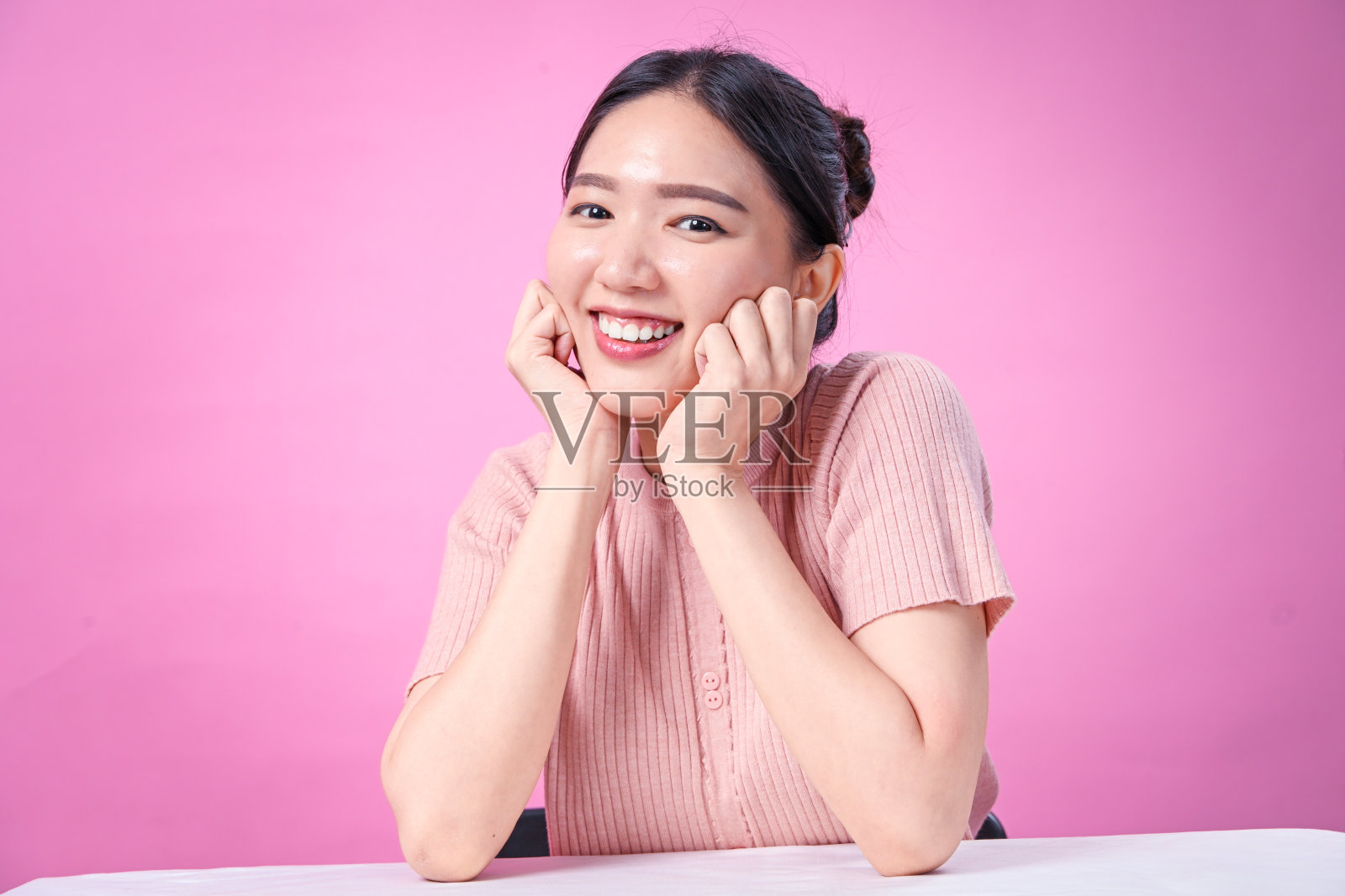 一位年轻的亚洲女性，深色头发扎起，穿着罗纹粉色短袖上衣，双手托腮，对着纯粉色背景露出灿烂的笑容，表情十分愉悦。照片摄影图片