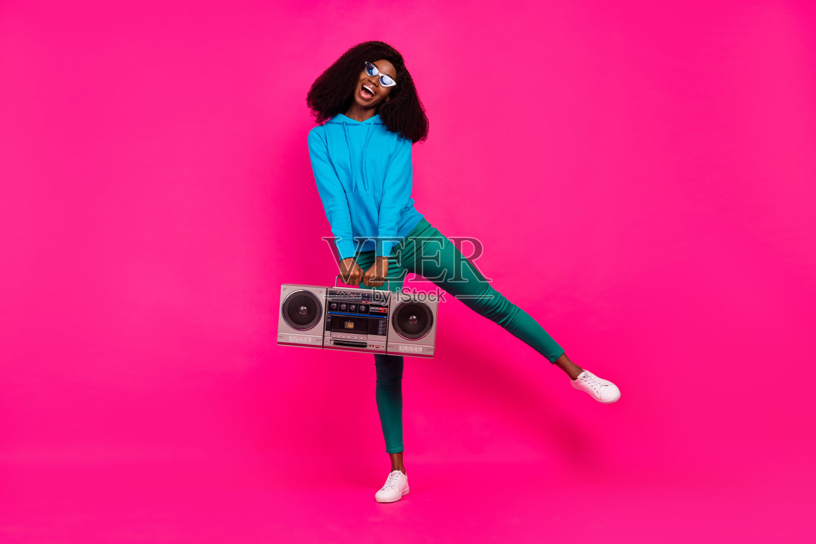 身穿蓝色运动衫的漂亮酷炫黑皮肤女子，在粉色背景前对着音响微笑跳舞照片摄影图片