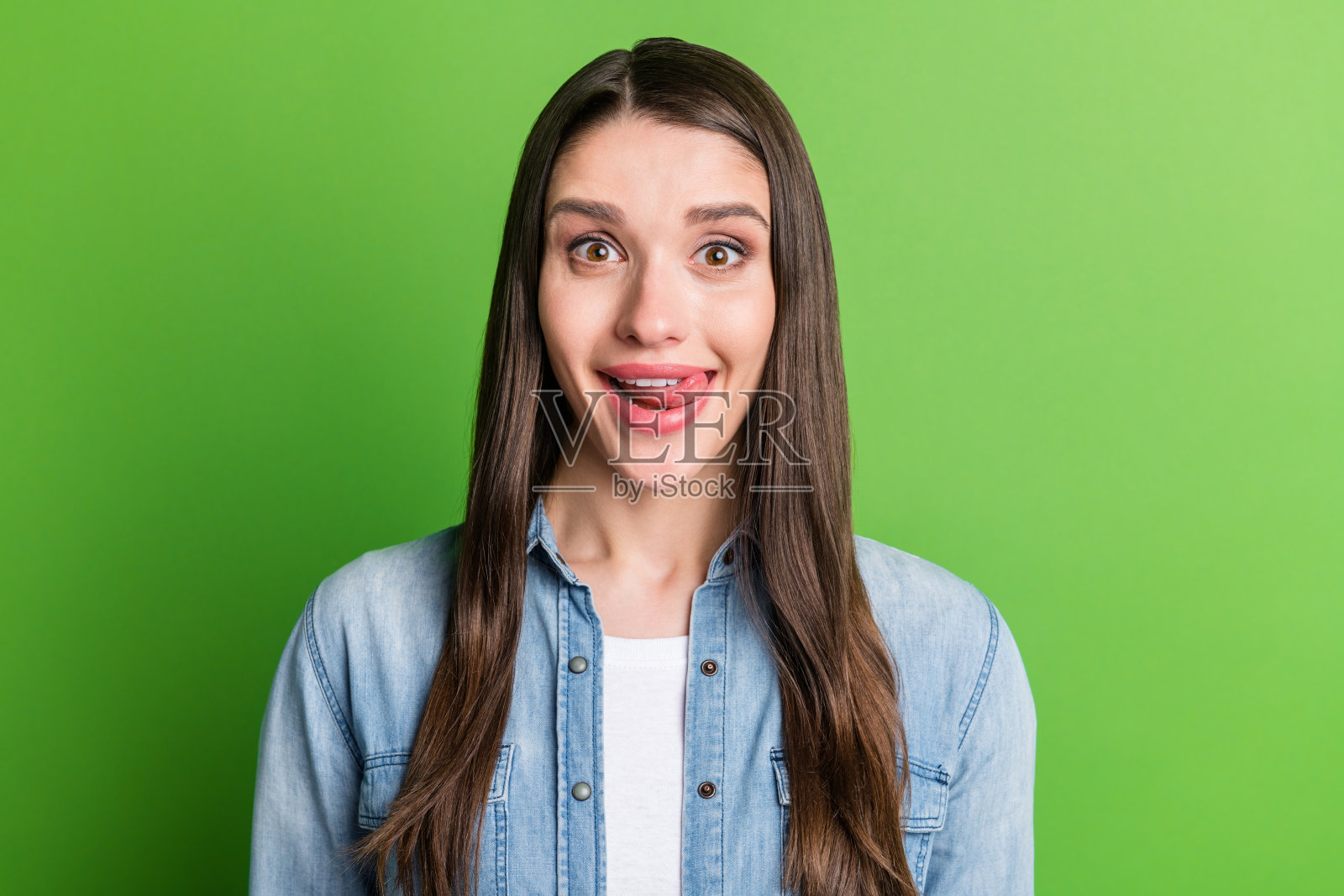 一位迷人的、时髦的、饥饿的、快乐的女孩，正舔着嘴唇，背景是明亮的绿色。照片摄影图片