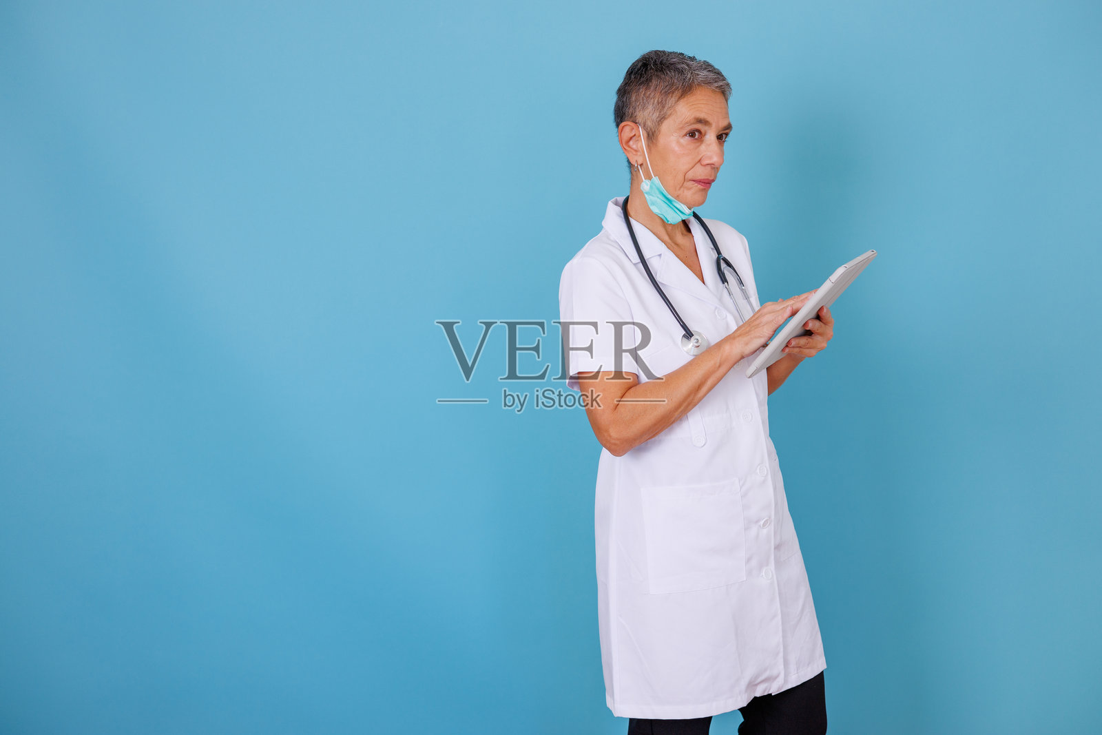 身穿白大褂、佩戴听诊器的经验丰富的医务人员在影棚内使用平板电脑查看患者信息照片摄影图片