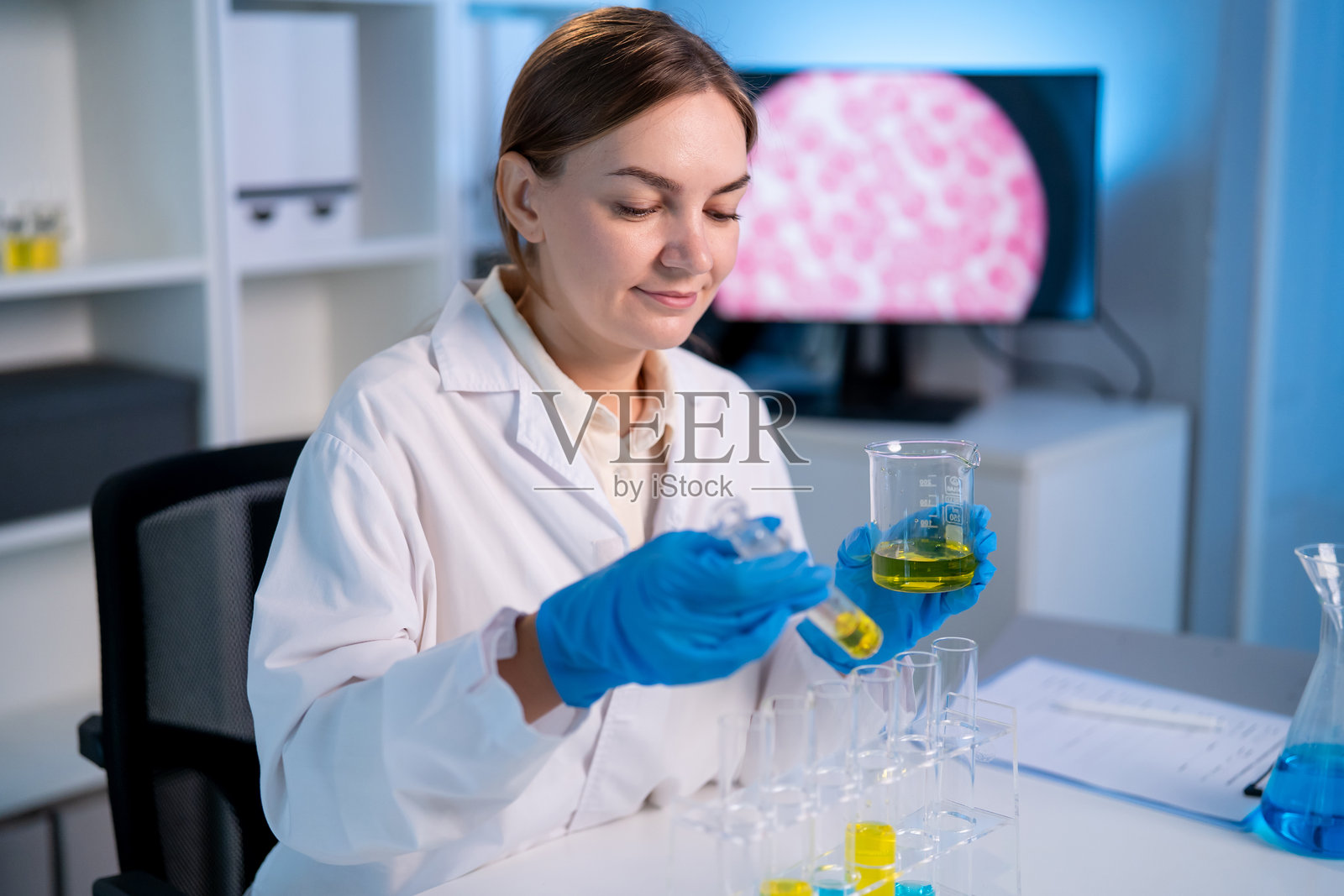 身穿白色实验服的高加索女科学家在实验室工作。一位富有魅力的女性技术员研究员坐在办公桌前，手持试管，为医学研发进行分析和测试。照片摄影图片