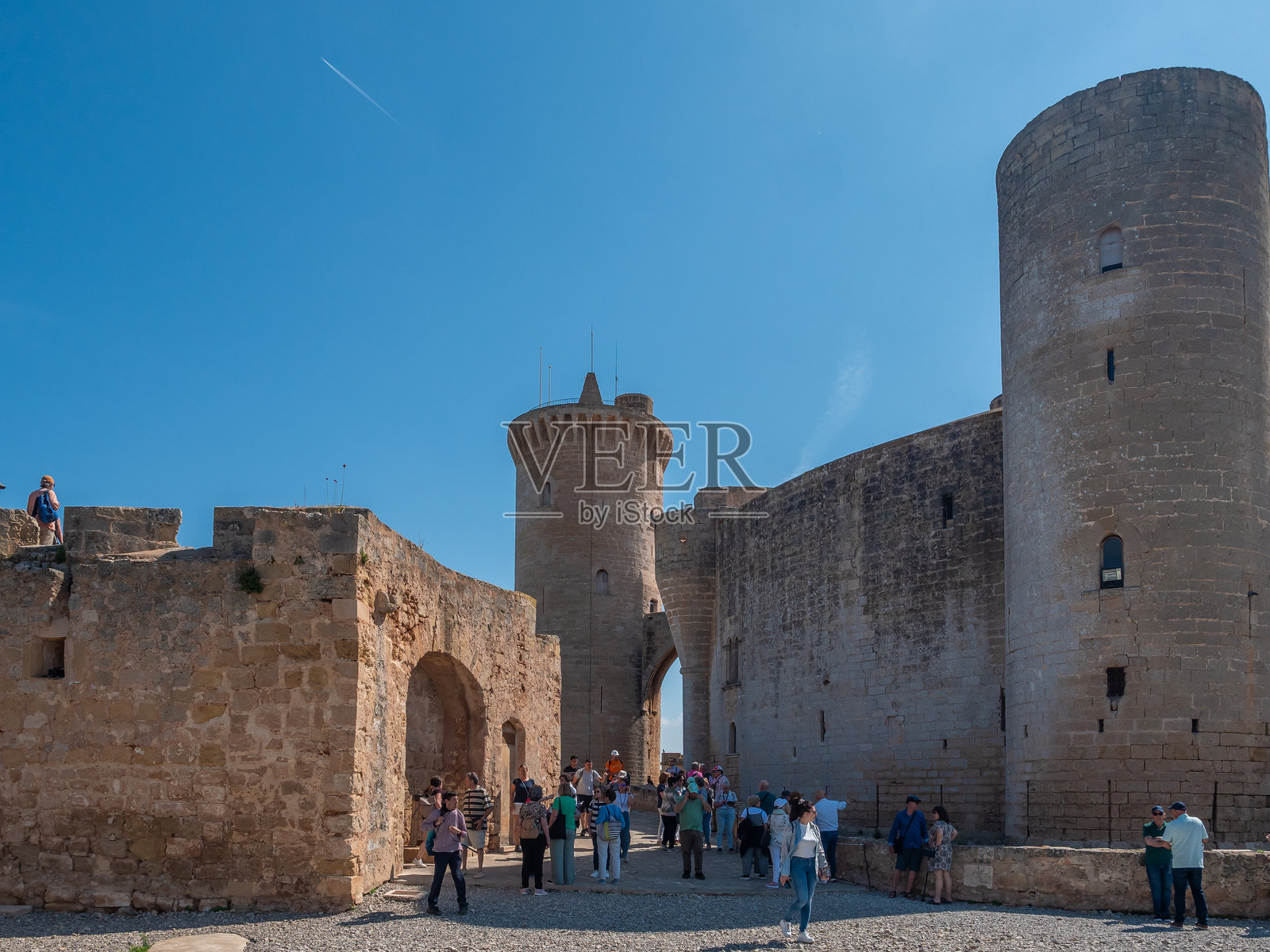 贝尔维尔城堡,帕尔马,一群游客站在一座古老的石头城堡前,城堡有一个突出的圆形塔楼,天空湛蓝,马略卡岛照片摄影图片