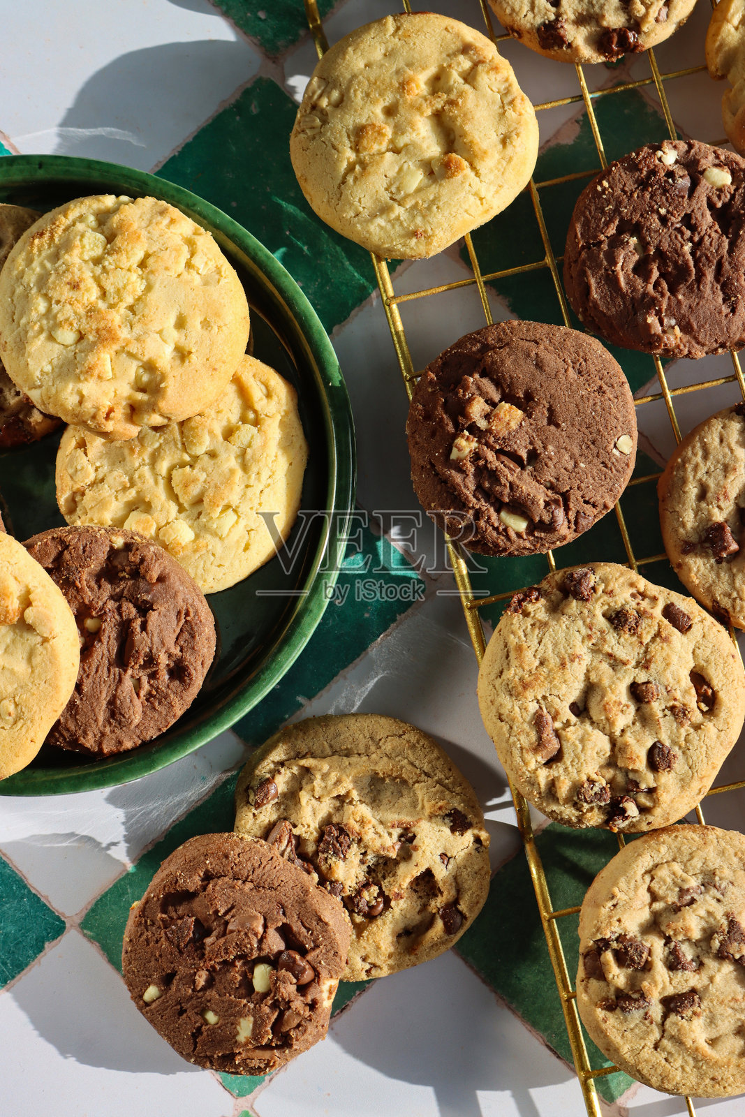 绿色盘子和金属冷却架上巧克力碎曲奇饼干圆的特写图片，节日三倍巧克力碎、牛奶巧克力碎和白巧克力碎饼干，白色和绿色格子图案背景，俯视角度照片摄影图片