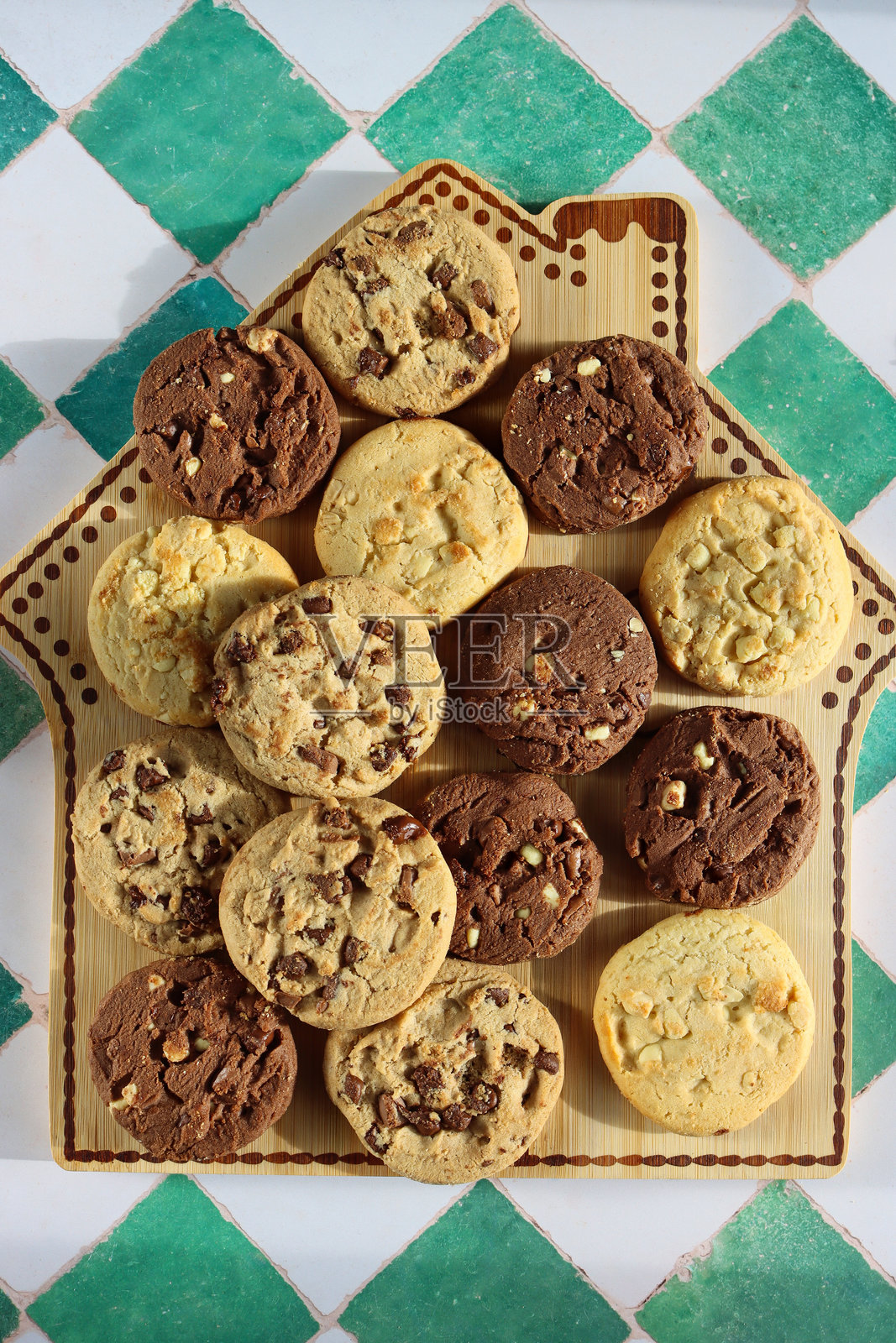 姜饼屋设计的面包板上摆放着巧克力碎饼干，节日氛围浓郁的 triple 巧克力碎、牛奶巧克力碎和白巧克力碎饼干，背景为白色和绿色格子图案，俯视角度。照片摄影图片
