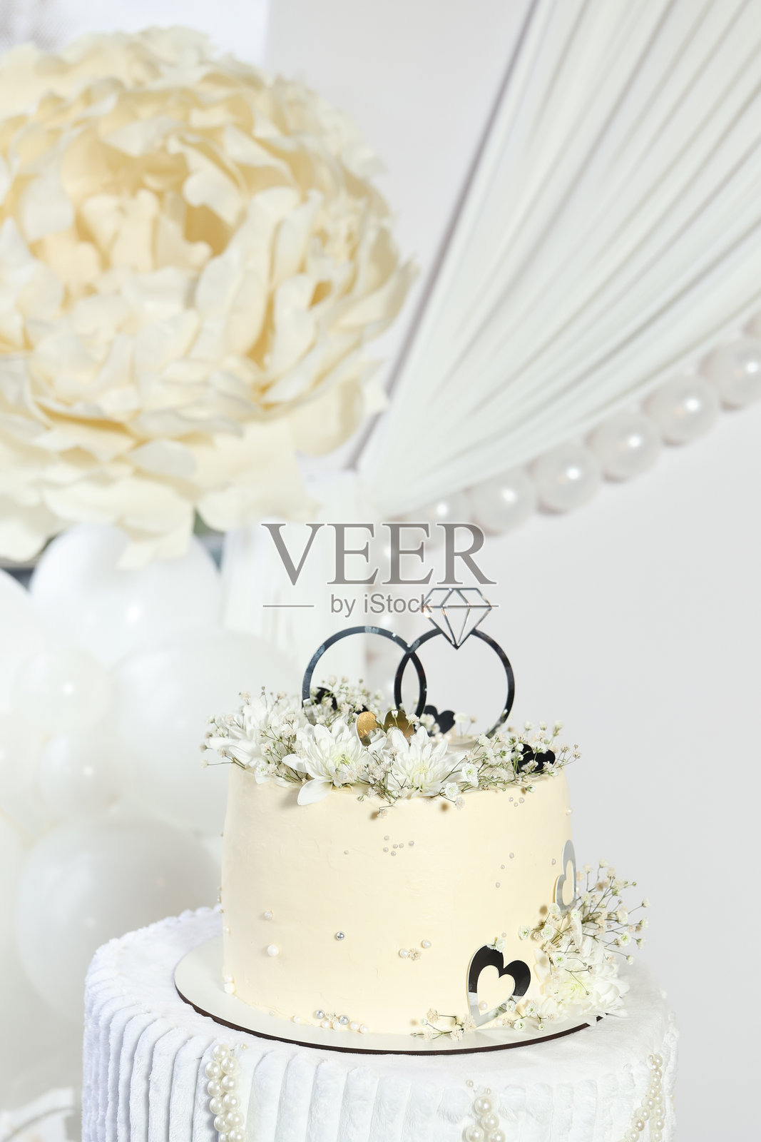 特写镜头：装饰着精致白色花朵和银色戒指顶饰的婚礼蛋糕，背景为超大花卉装饰和优雅垂坠的织物。柔和的色调营造出精致的婚礼氛围。照片摄影图片