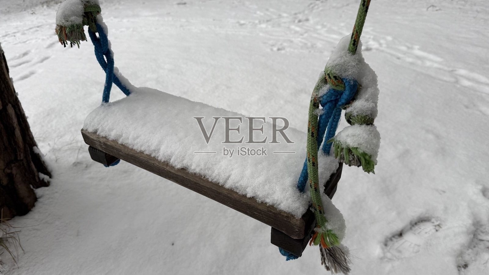 冬季森林中覆盖着新鲜积雪的木制秋千特写——废弃的户外游乐场场景，带有彩色的绳索和雪地上的脚印照片摄影图片