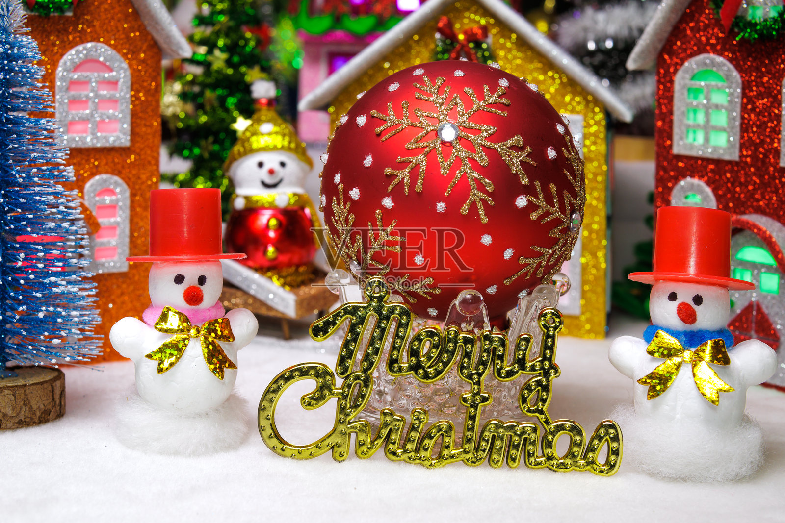 在圣诞村的房屋之间，有一个坐在雪橇上的雪人，还有两个戴着红帽子的雪人，中间放着一个红色的圣诞装饰球和字母。照片摄影图片