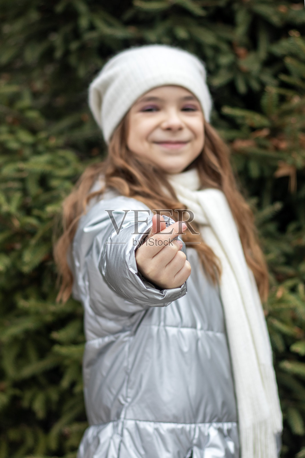年轻女孩穿着银色羽绒服和白色绒线帽，在户外对着镜头微笑并举起手指照片摄影图片