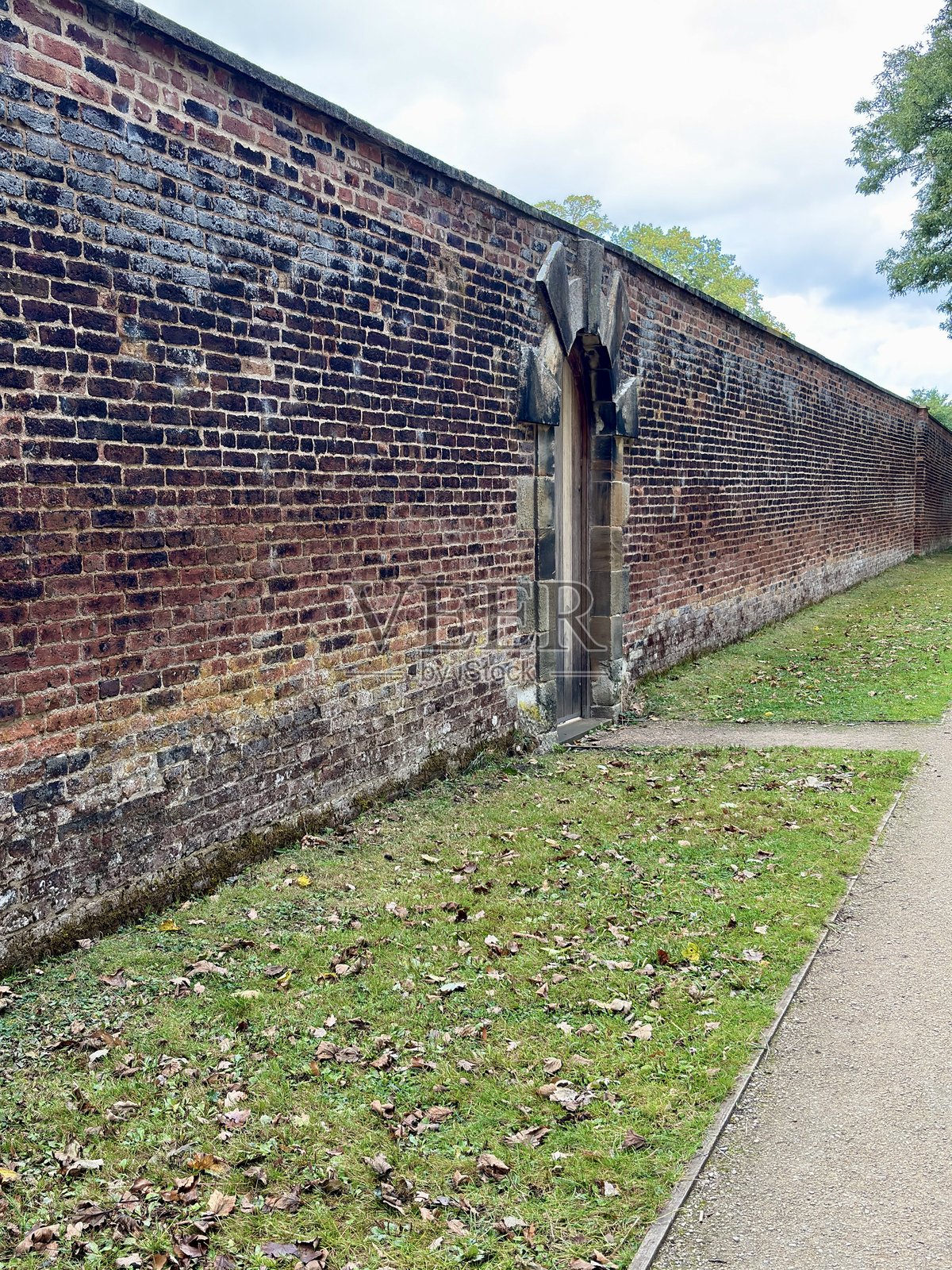 一条安静小路旁矗立着一堵长长的砖墙，墙上有一个木门。前景的草地为这幅宁静、略带忧郁的户外场景增添了质感。照片摄影图片