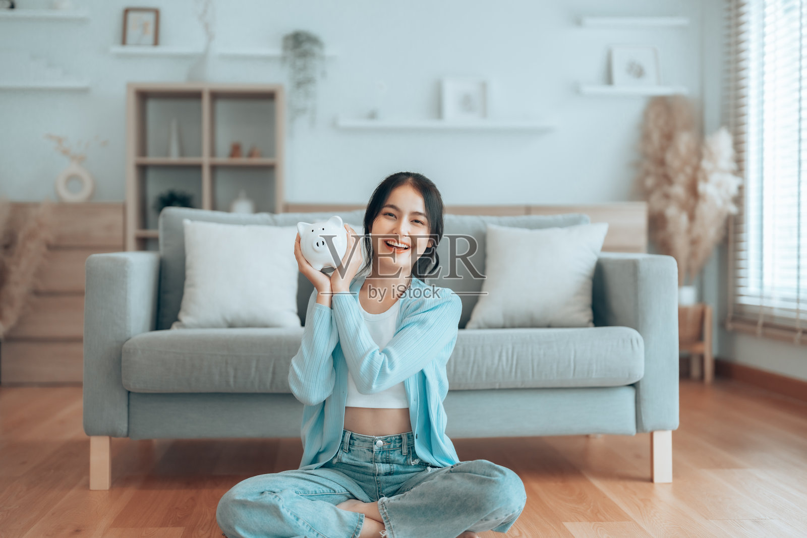 年轻的亚洲女性在家中坐在地板上，手里拿着存钱罐，寓意储蓄和财务投资。照片摄影图片