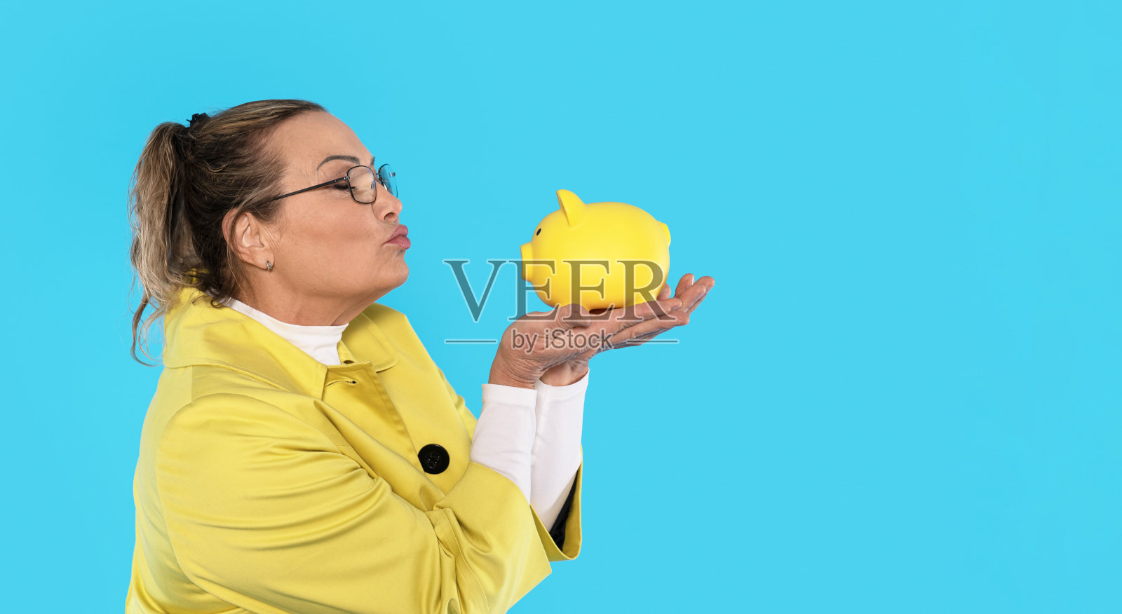 女子身穿黄色外套，在明亮背景前亲吻黄色储蓄罐照片摄影图片