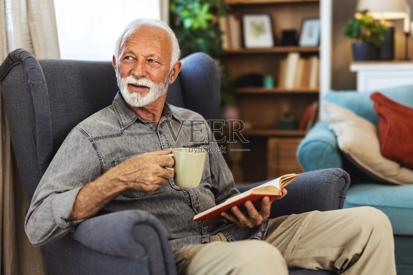 一位老人正在家中阅读书籍、喝咖啡，享受退休或宁静的客厅时光，手捧小说和饮品。一位平静的老年人，带着热饮、茶和读物，在休息室里汲取知识。照片摄影图片