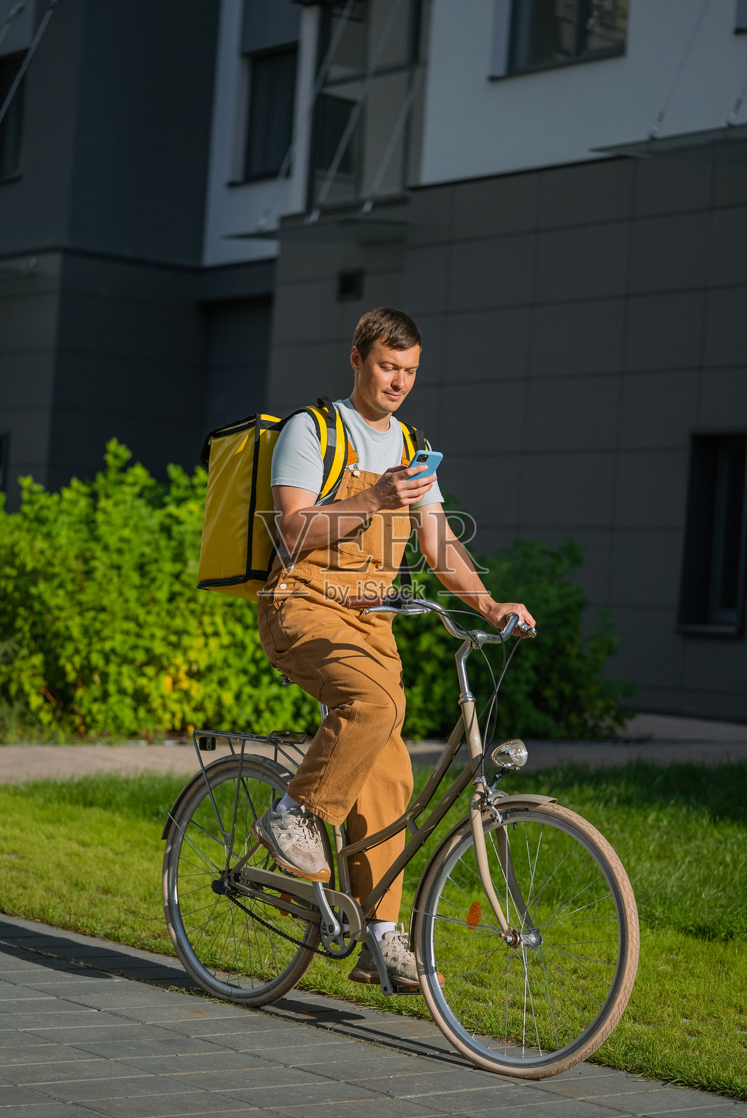 穿着工装裤和黄色配送背包的自行车快递员，在现代城市街区骑行，一边看着智能手机屏幕导航。照片摄影图片