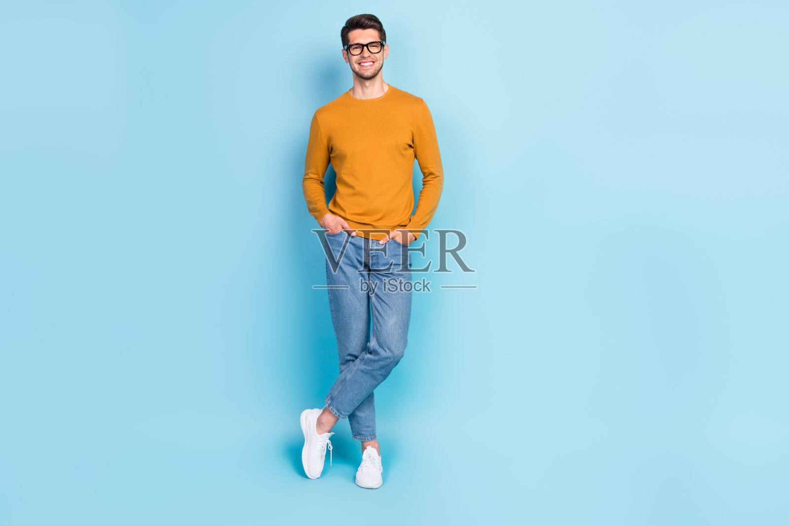 身穿黄色套头衫、戴着眼镜、双手插在口袋里、面带微笑的可爱自信的年轻男性，在蓝色背景前照片摄影图片