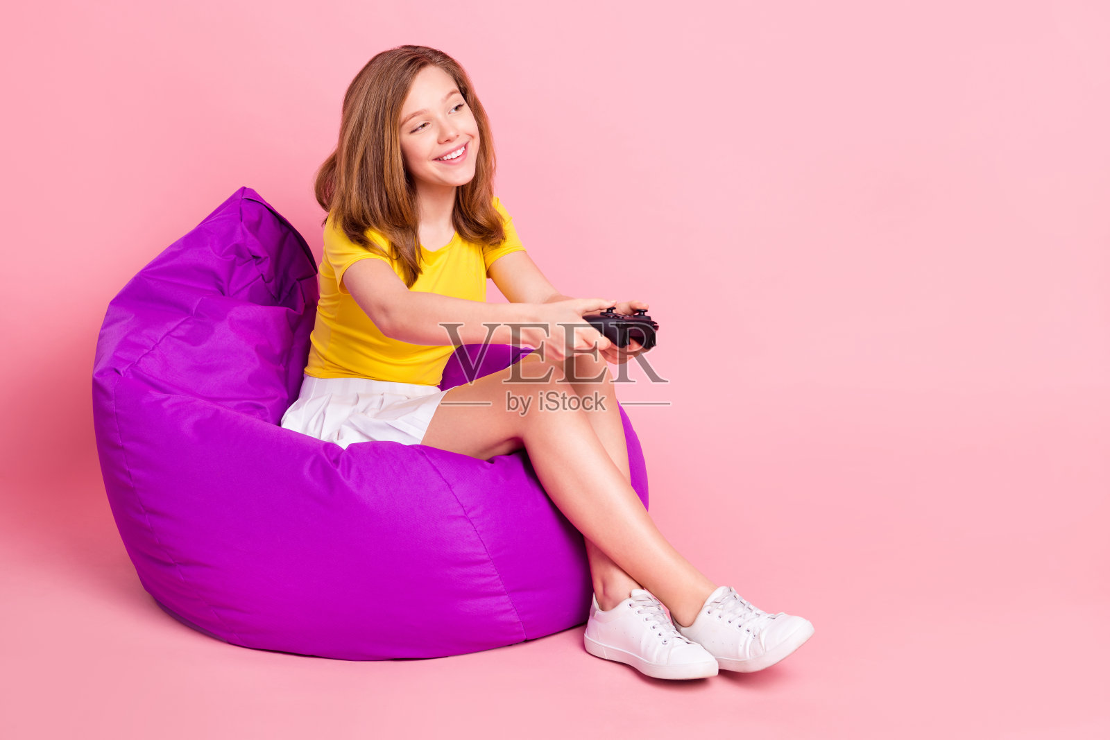 年轻女孩坐在豆袋椅上，手持游戏手柄，开心地玩着电子游戏，背景为柔和的彩色渐变，全身侧面照照片摄影图片