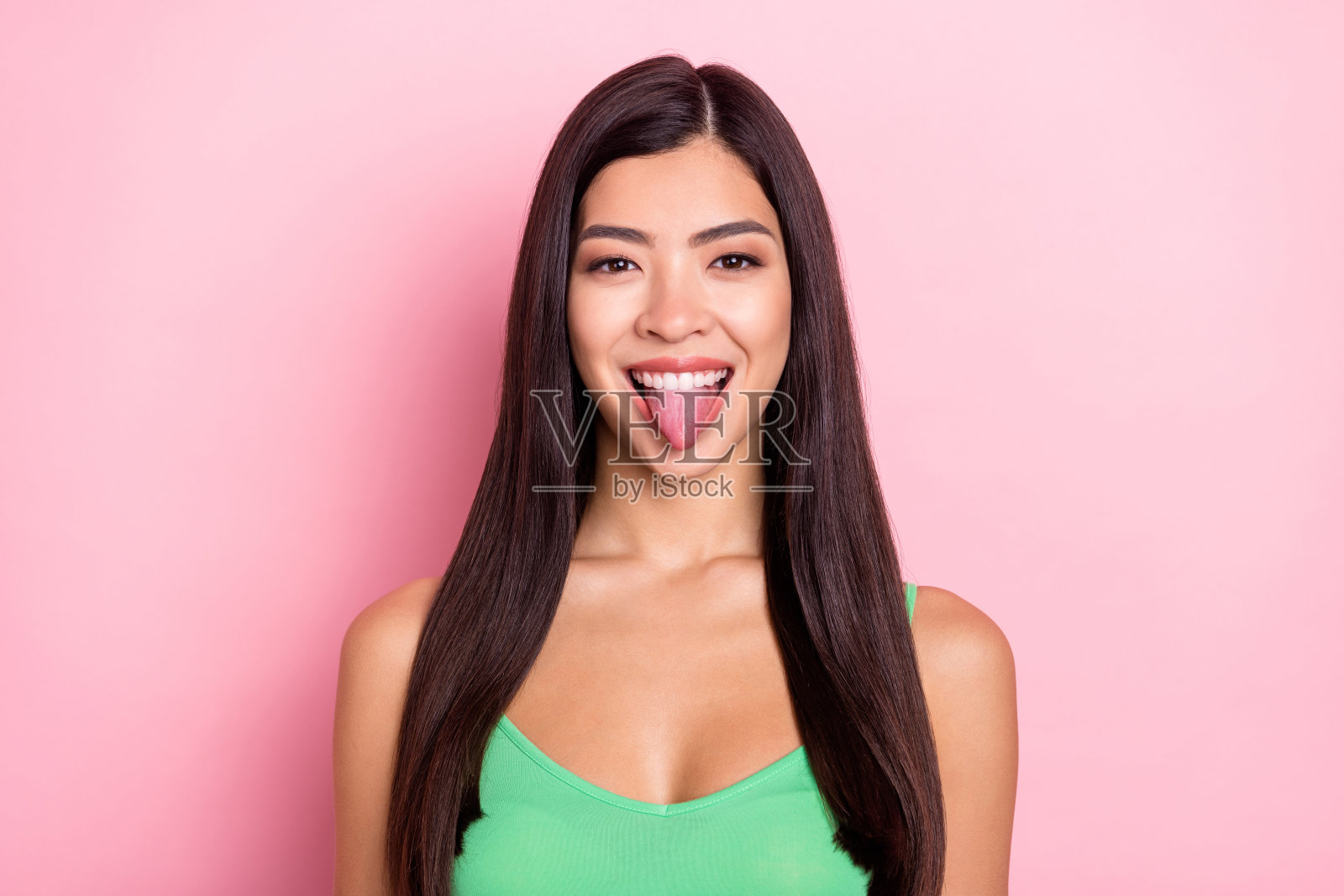 穿着绿色背心的甜美孩童般年轻女子，面带笑容，伸出舌头，在粉色背景下拍摄照片摄影图片
