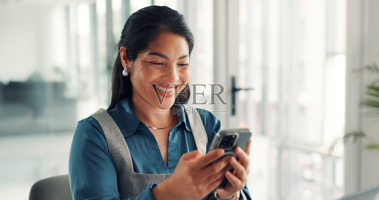 办公室里，一位快乐的女商人在使用手机进行网络连接、通讯或邮件应用的操作。她面带微笑，运用科技，这位女性正在使用手机进行在线联系、发短信或在线研究。照片摄影图片