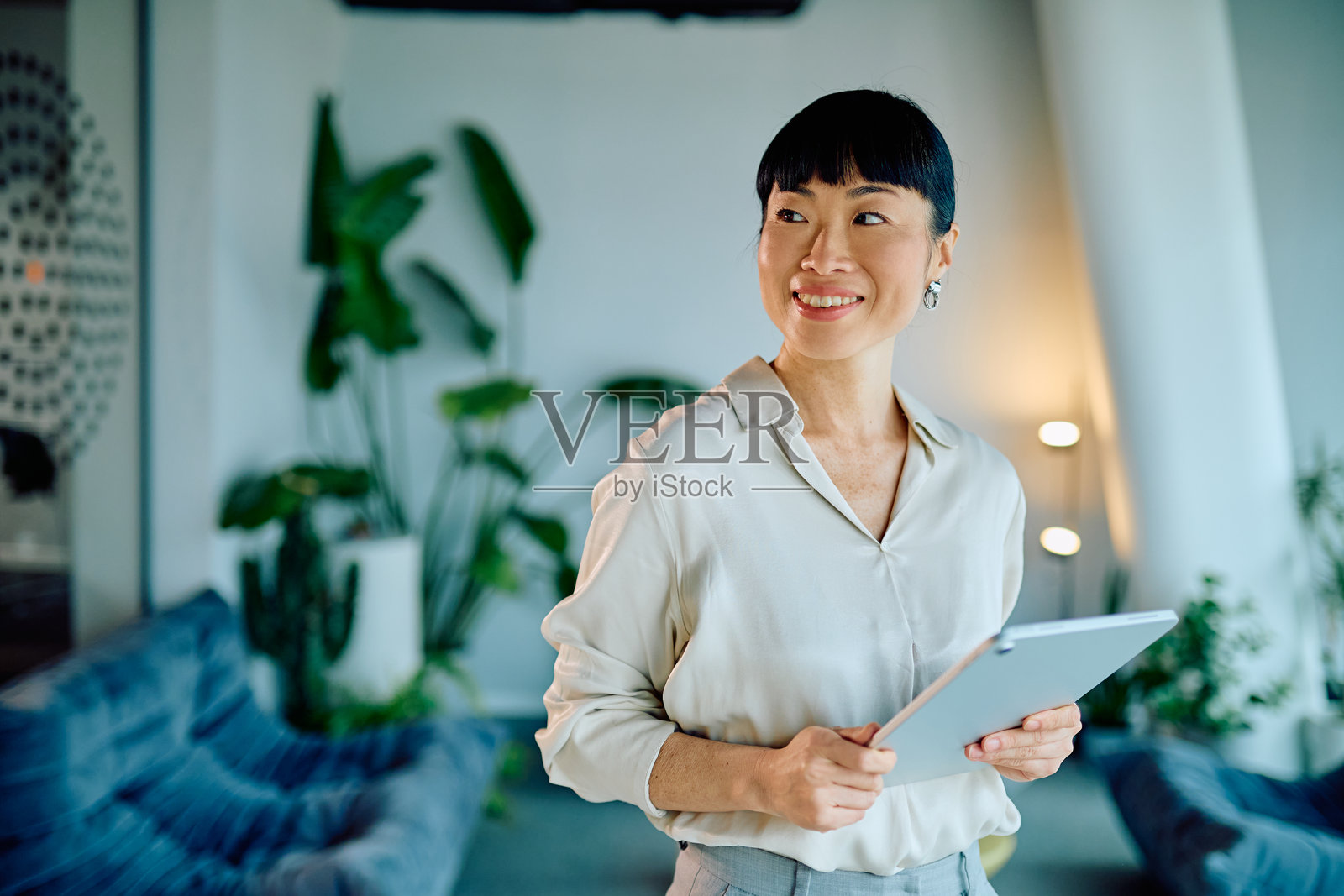 一位亚洲女商人微笑着，目光望向别处，手里拿着平板电脑，站在现代办公室里照片摄影图片