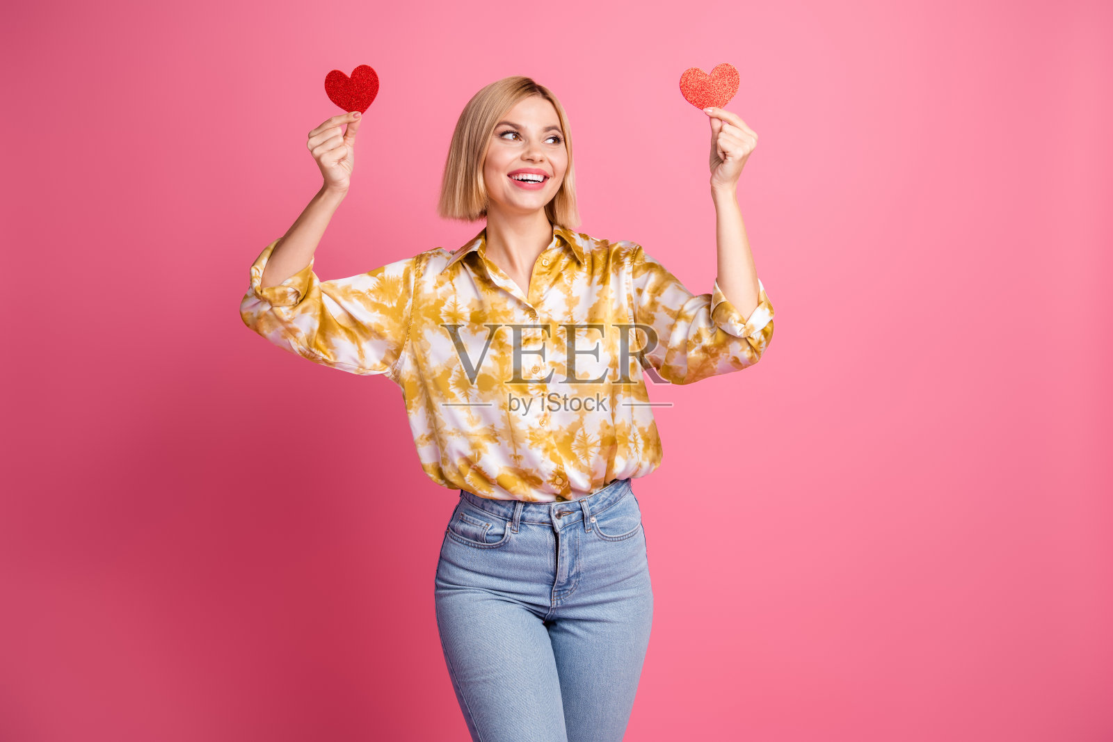 一位金发波波头的年轻女子，身穿黄色衬衫，手持两个闪光的心形，在粉色背景前庆祝爱情与时尚。照片摄影图片