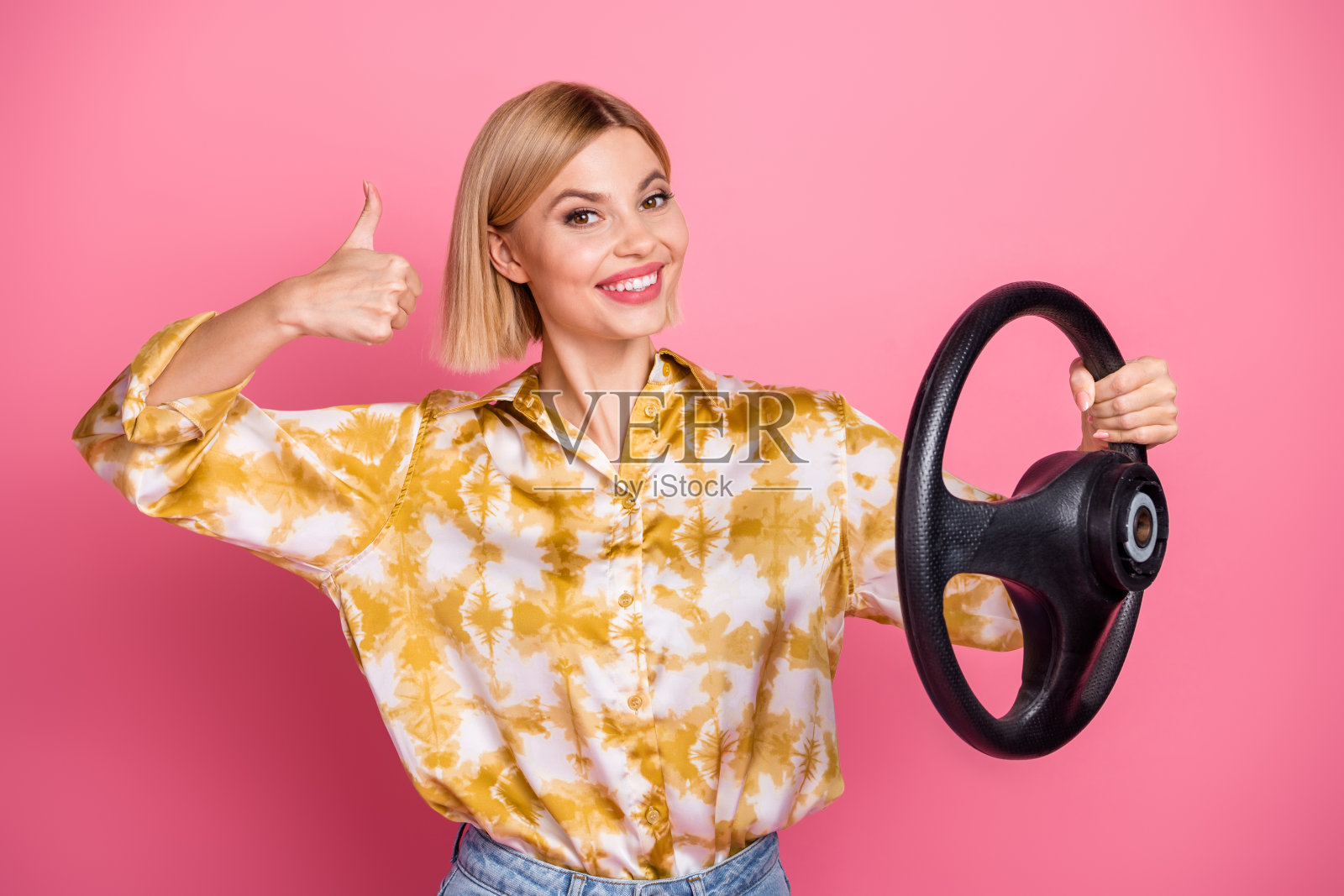 身穿印花黄衬衫的年轻女子手握方向盘，在粉色背景前微笑照片摄影图片