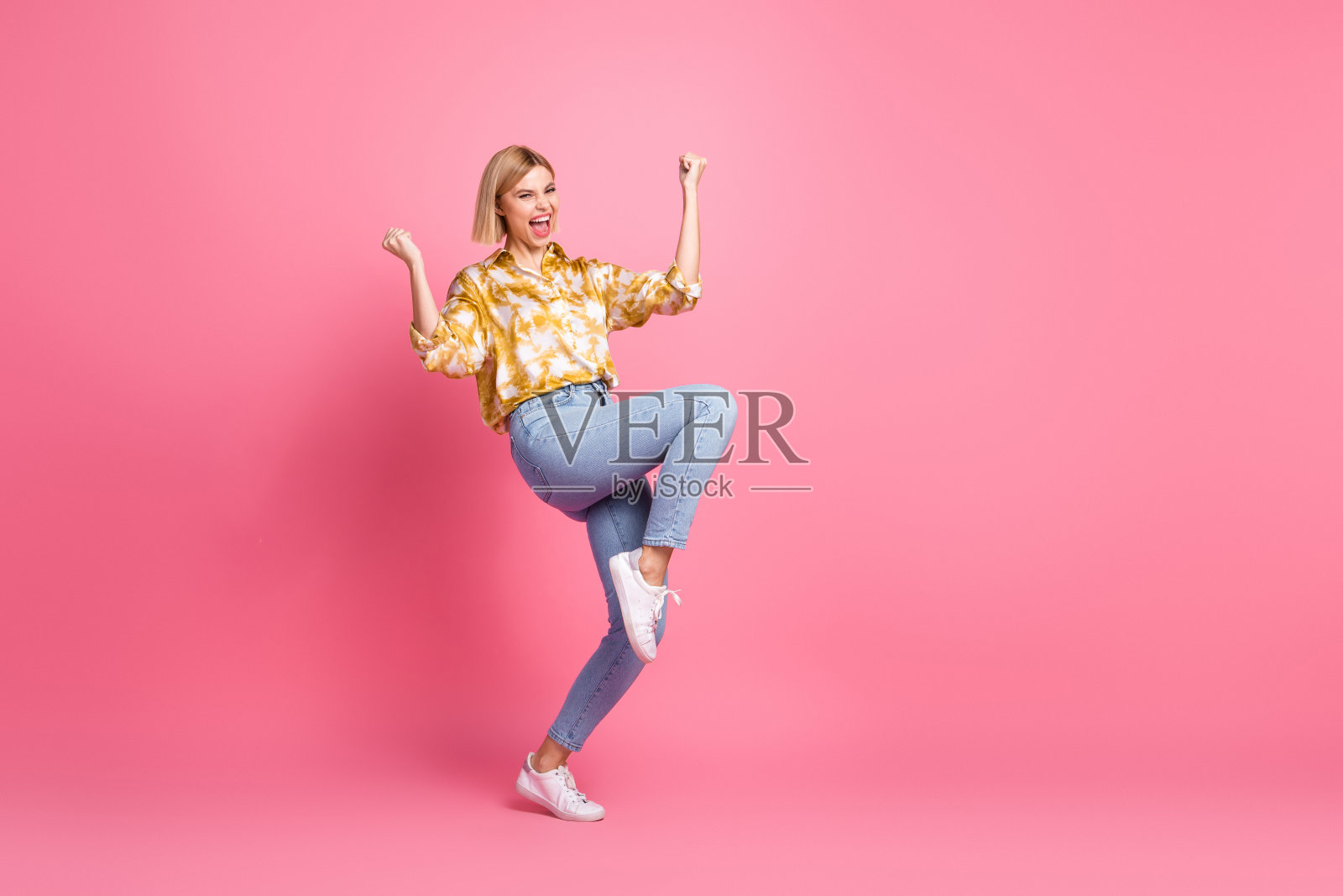 身穿黄色上衣和牛仔裤的年轻女子在粉色背景前欣喜雀跃，用于生活时尚广告照片摄影图片