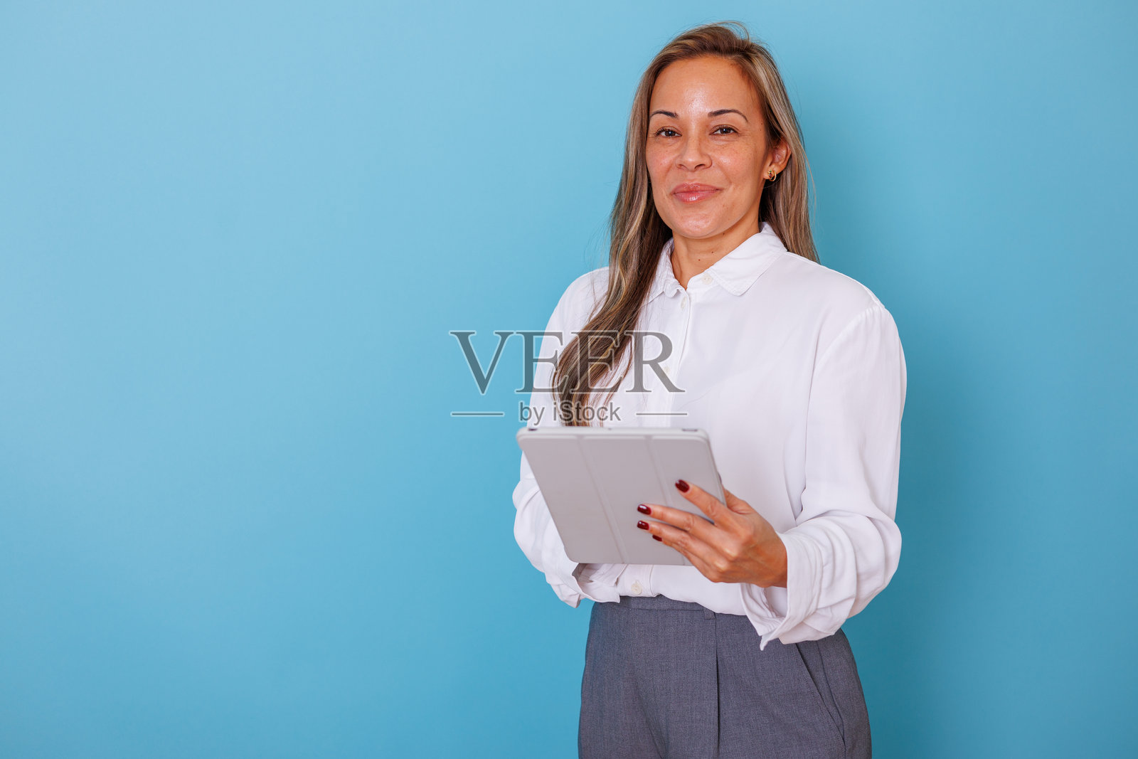一位自信的成熟的女性，面带平静的表情，手持数字平板电脑，站在蓝色背景前。照片摄影图片