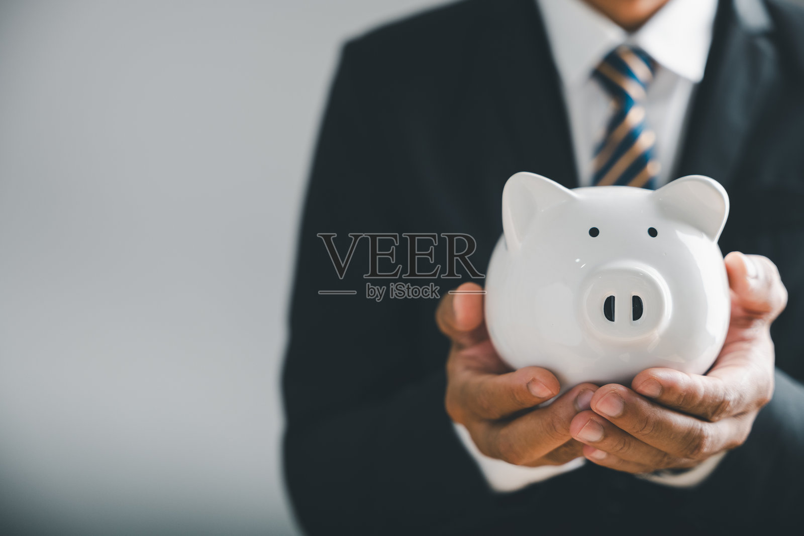 西装革履的商人双手握着一个白色的储蓄罐，背景是模糊的灰色墙壁。照片摄影图片