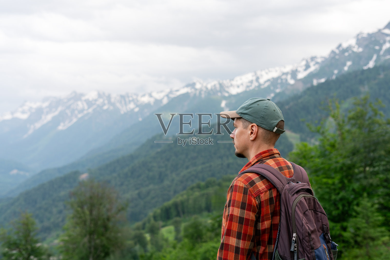 背包客在山顶观景点侧身而立，眺望远方覆盖着森林的山丘和雪山。概念为徒步、探险旅行，可供复制文字的空间。照片摄影图片