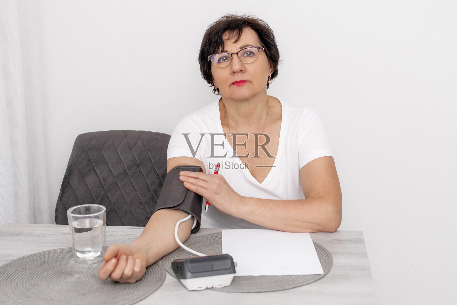 一位老年女性在家中使用电子血压计测量血压，她坐在摆有水杯和笔记本的桌子旁，在日常环境中监测健康和心血管状况照片摄影图片