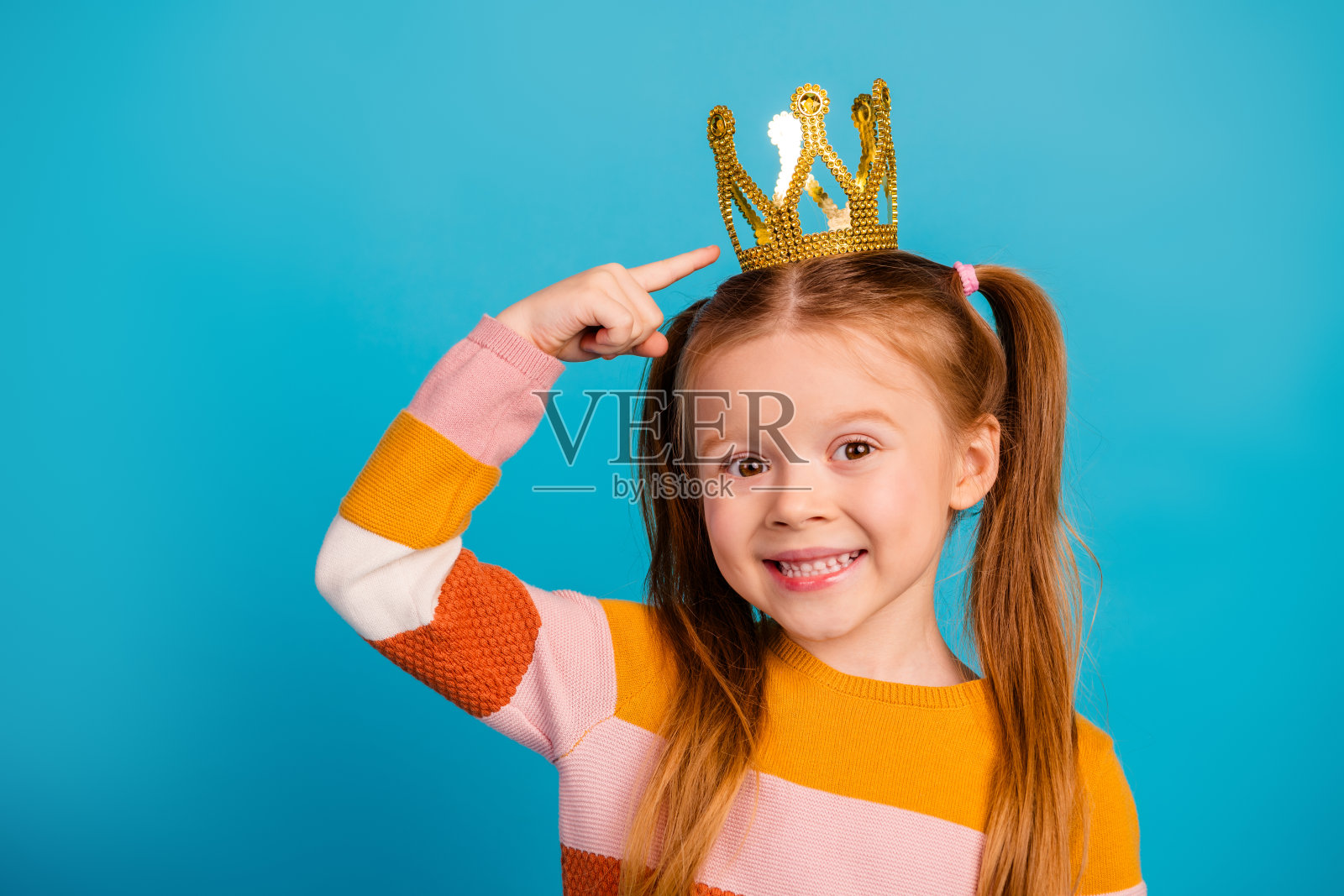 一个快乐的小女孩戴着金色皇冠，在明亮的蓝色背景前带着俏皮的笑容，指向自己的皇冠，画面充满欢乐和玩趣。照片摄影图片