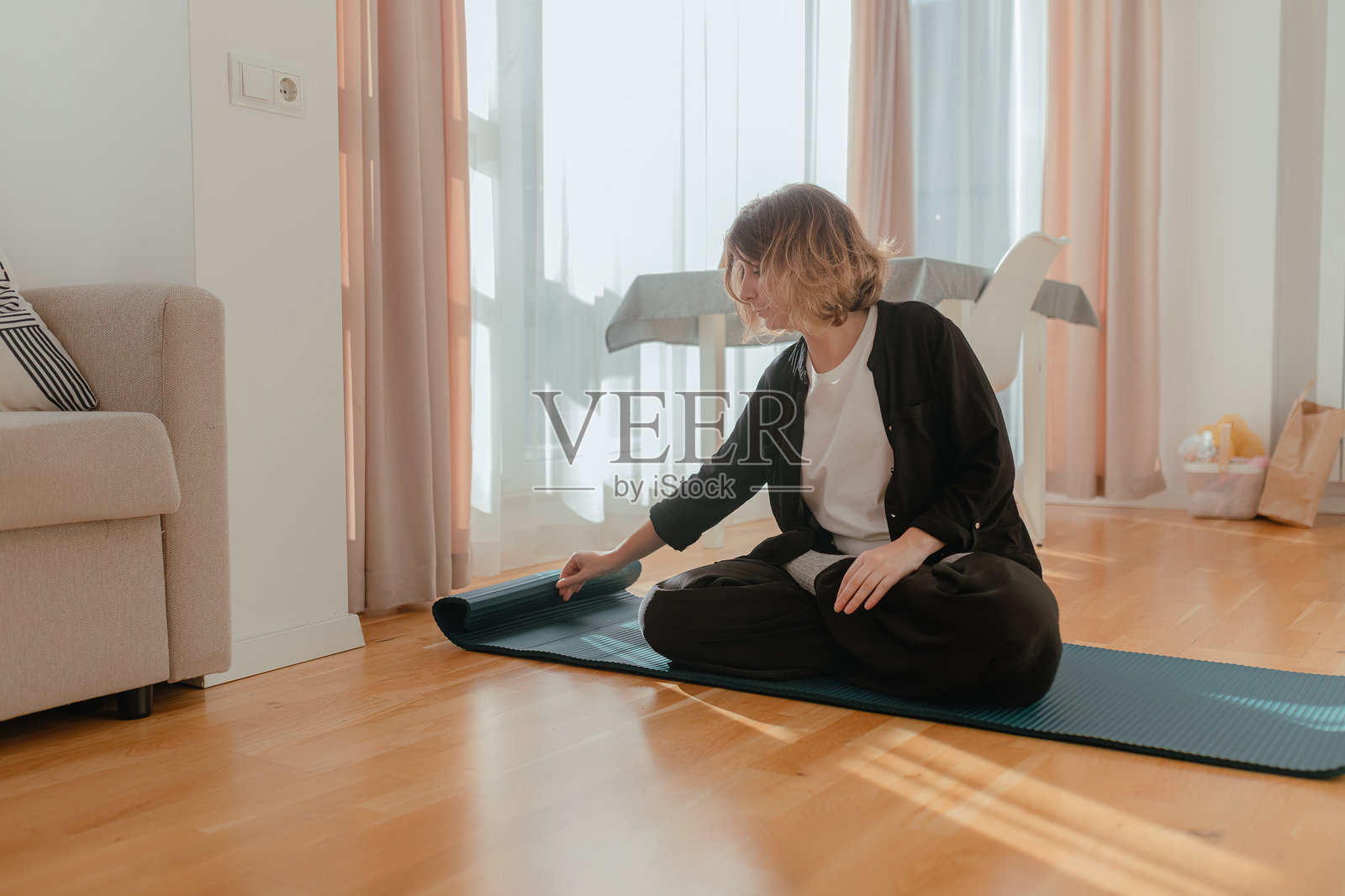 一位女士盘腿而坐，在明亮的室内阳光透过木地板，调整着卷起的瑜伽垫。照片摄影图片