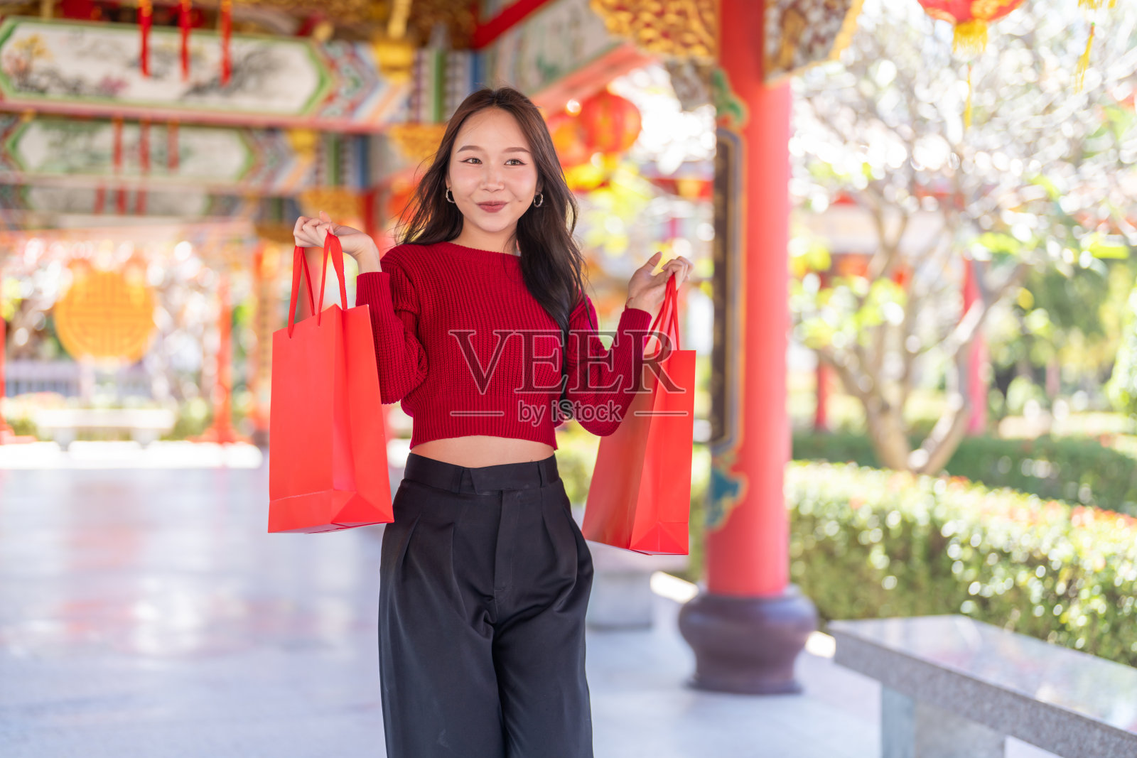 一位亚洲女孩在中国寺庙里开心地拿着红色购物袋，充满喜悦地庆祝农历新年，融合了节日传统、现代购物乐趣、中国新年购物和节日送礼的欢乐时光。照片摄影图片