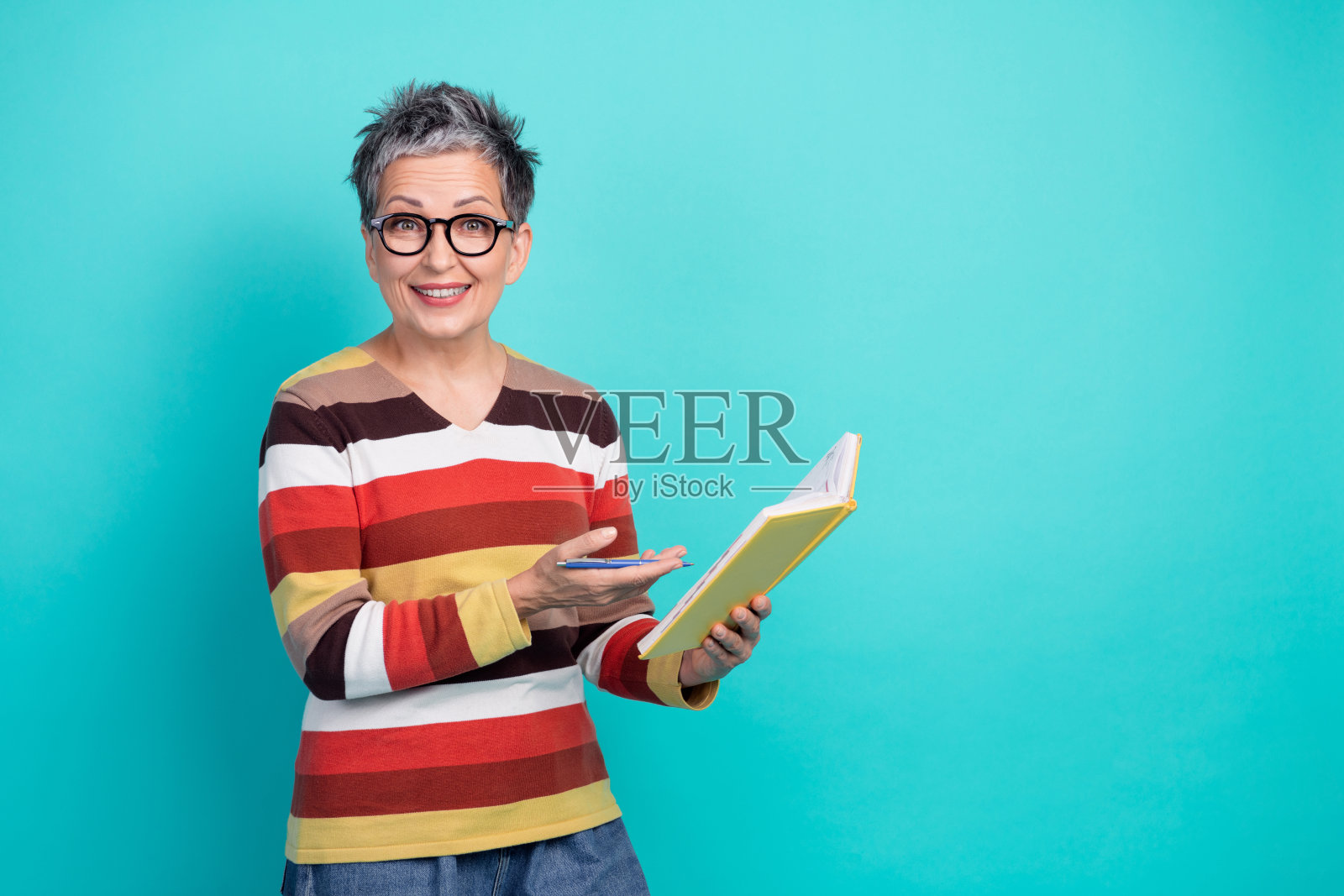 一位留着短发、戴着眼镜、穿着条纹毛衣的成熟的女性，手持笔记本和笔，微笑着看向镜头，背景为蓝色，为肖像照照片摄影图片