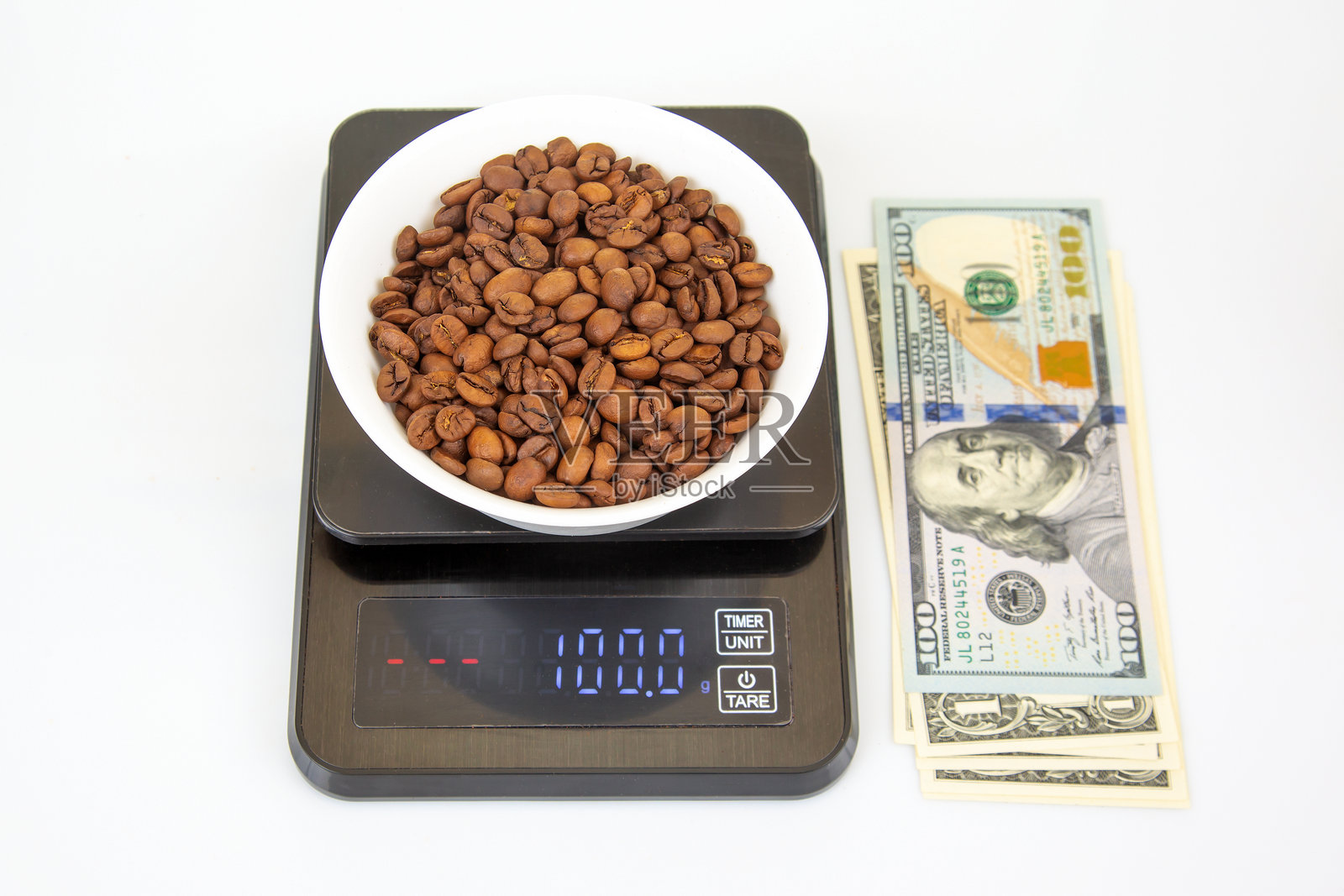 一个装满烘焙咖啡豆的白色碗放在电子秤上，显示100克，旁边放着美元钞票，背景为白色。照片摄影图片