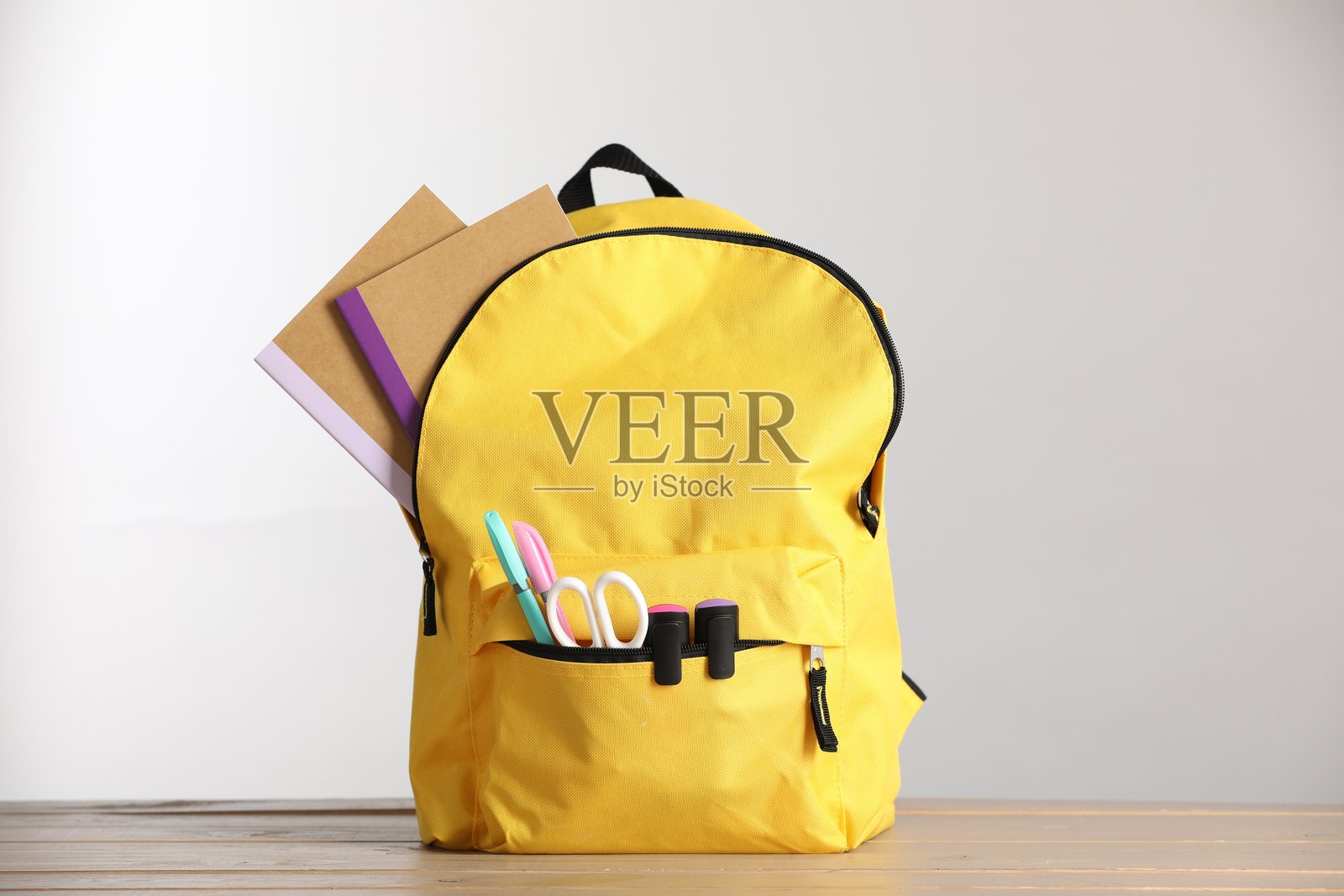 黄色的背包和各种文具放在木桌上，背景为浅色。照片摄影图片