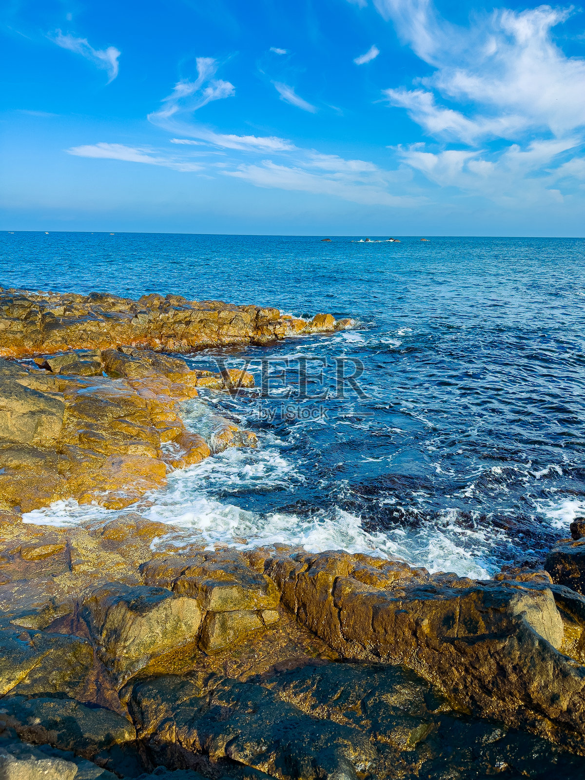 在明亮的白昼，海浪拍打着岩石海岸，蓝天下的背景中有远处的船只照片摄影图片
