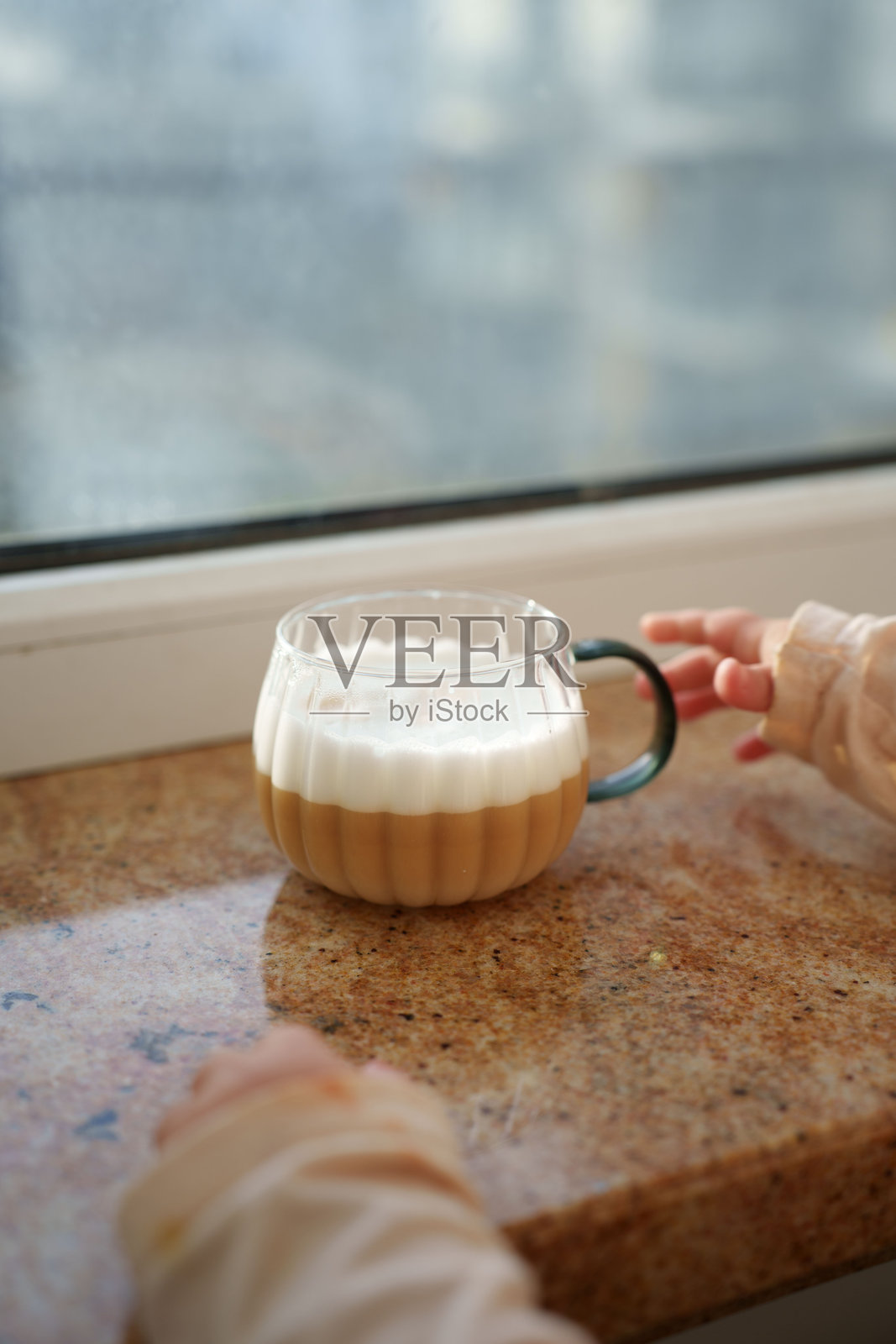阳光透过窗户洒在光滑的大理石台面上，一杯带有厚实手柄的透明杯子里的拿铁咖啡冒着绵密的泡沫，一双小手带着好奇而温柔地伸了过来，室内，日间，咖啡因照片摄影图片