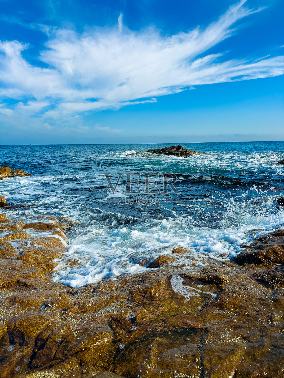 白昼时分，海边海岸地区的岩石海岸上，海浪拍打着，海天交接处是蓝天白云。照片摄影图片