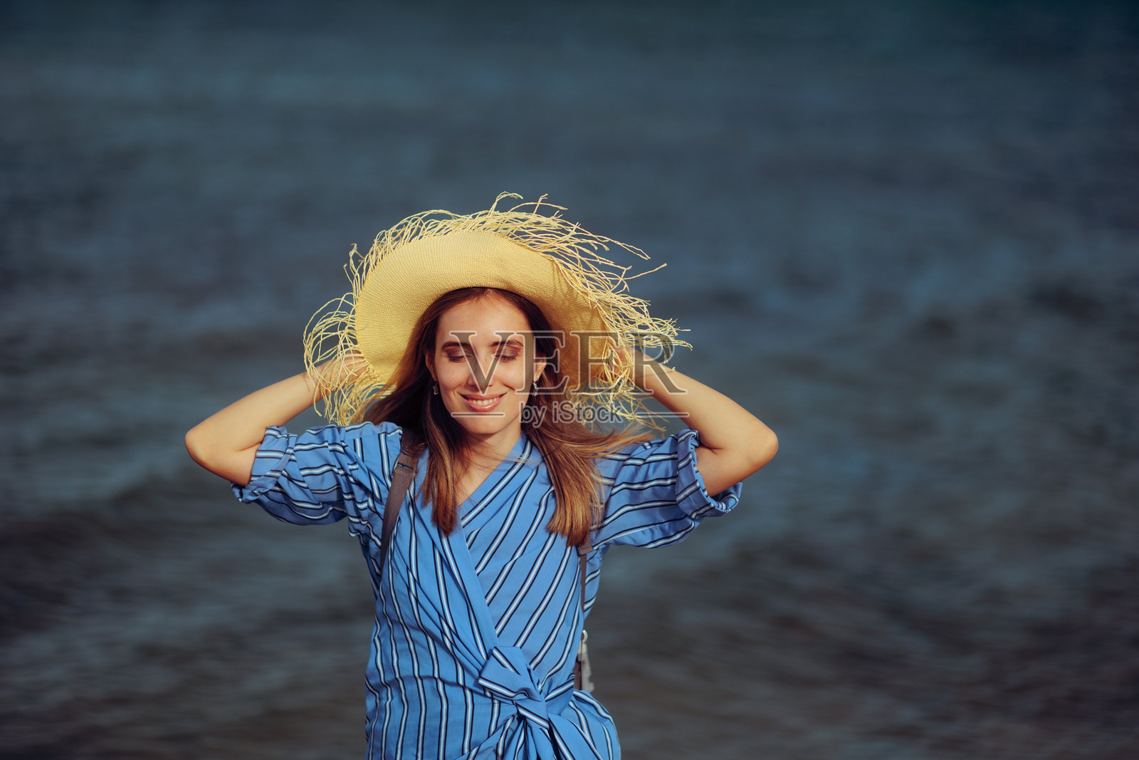 戴着草帽、穿着蓝色连衣裙、在海边的快乐女人照片摄影图片