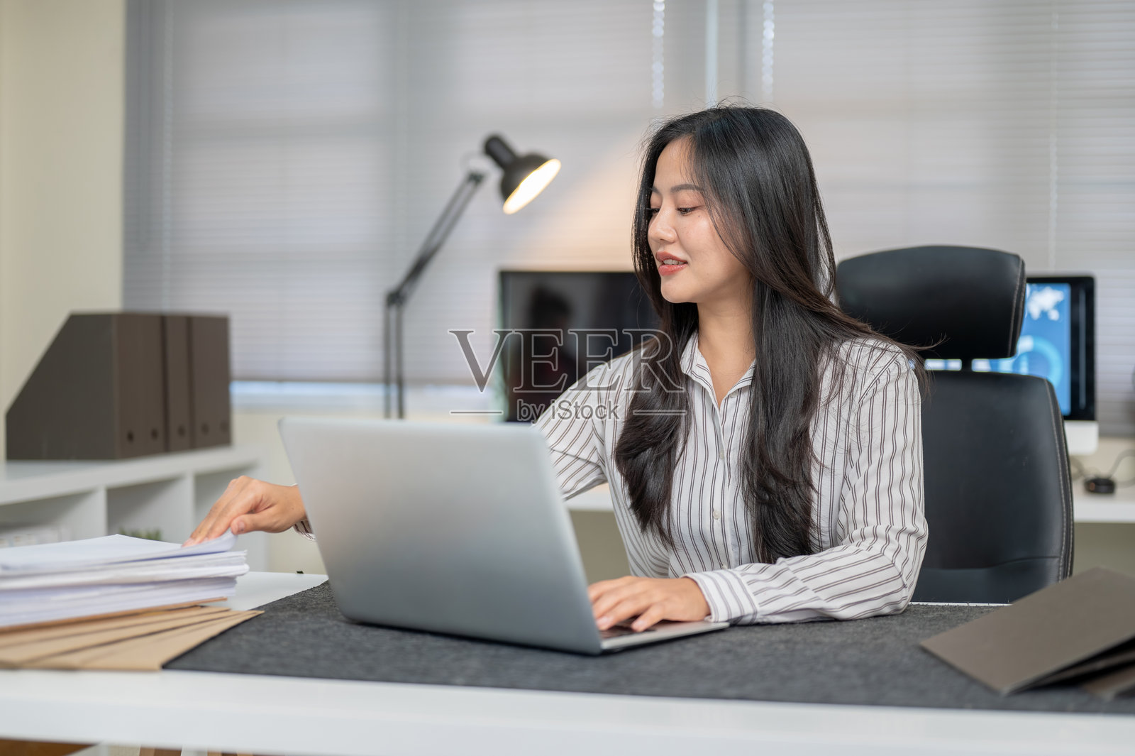 穿着条纹衬衫的漂亮亚洲女商人，在桌边笔记本电脑旁，抽出文件。照片摄影图片