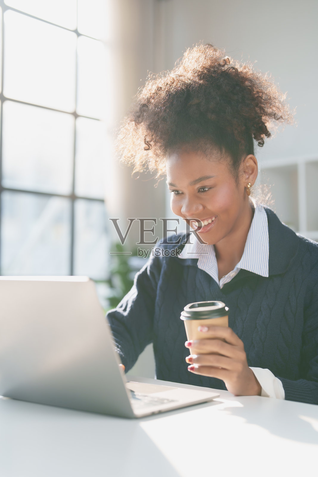 一位年轻的黑人女商人一边微笑着看着笔记本电脑，一边拿着咖啡杯，在一个阳光明媚的现代办公室里享受着高效的远程工作日照片摄影图片