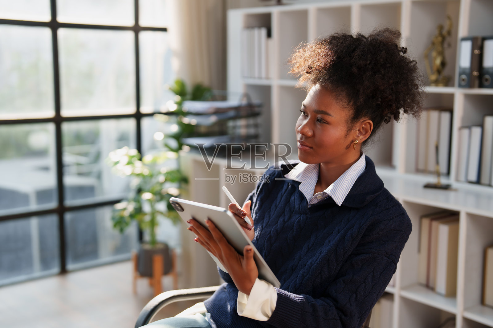 一位年轻的黑人女性在家里的办公室，手持数位板和触控笔，在现代、阳光充足的工作空间里，她正在深入思考，规划和创作数字内容。照片摄影图片