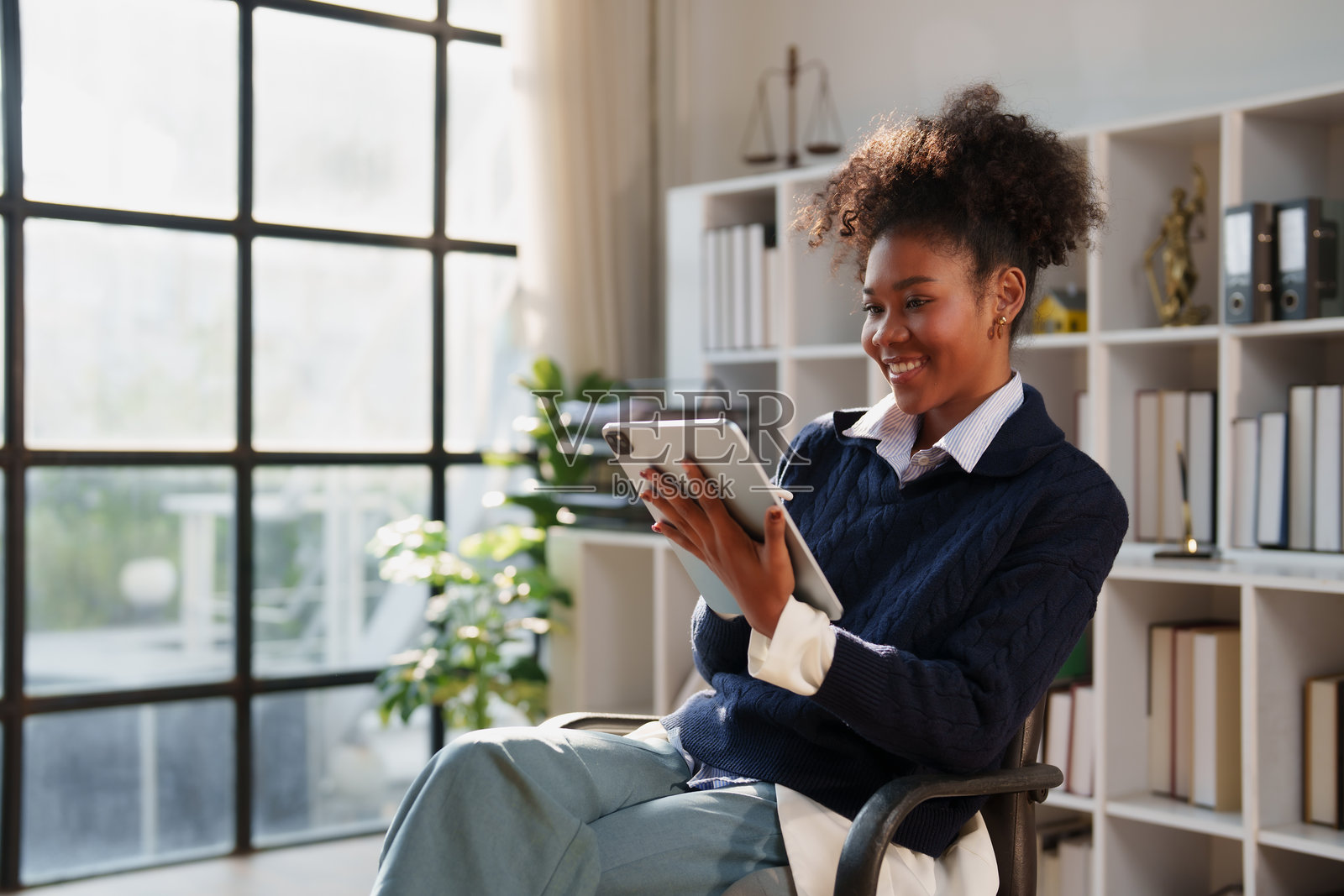 一位非裔美国女律师在现代律师事务所的办公室环境中，一边微笑着使用数字平板电脑工作，一边处理法律文件和客户案件。照片摄影图片